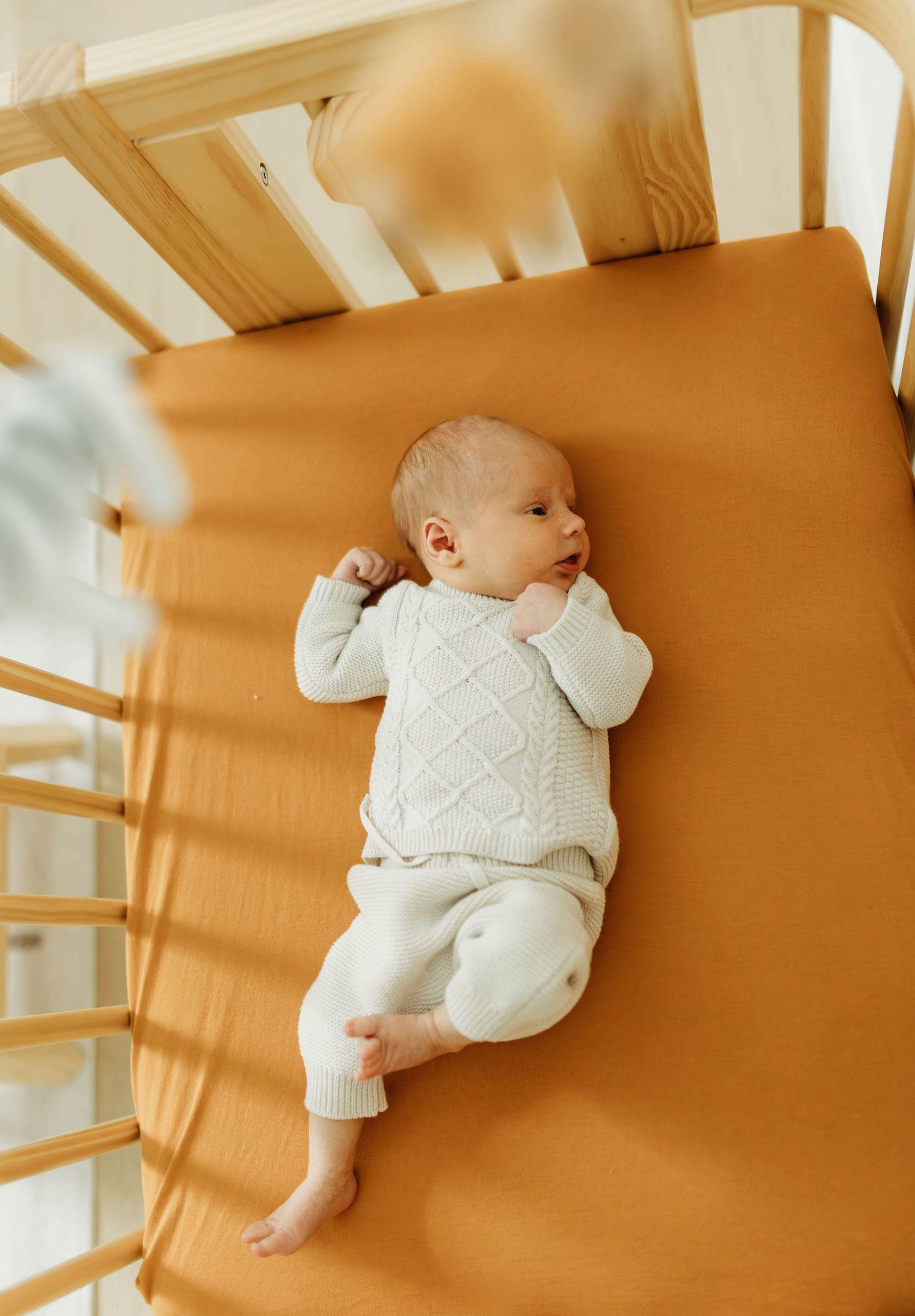 Newborn baby in white knit outfit lying awake in wooden crib with caramel sheet.
