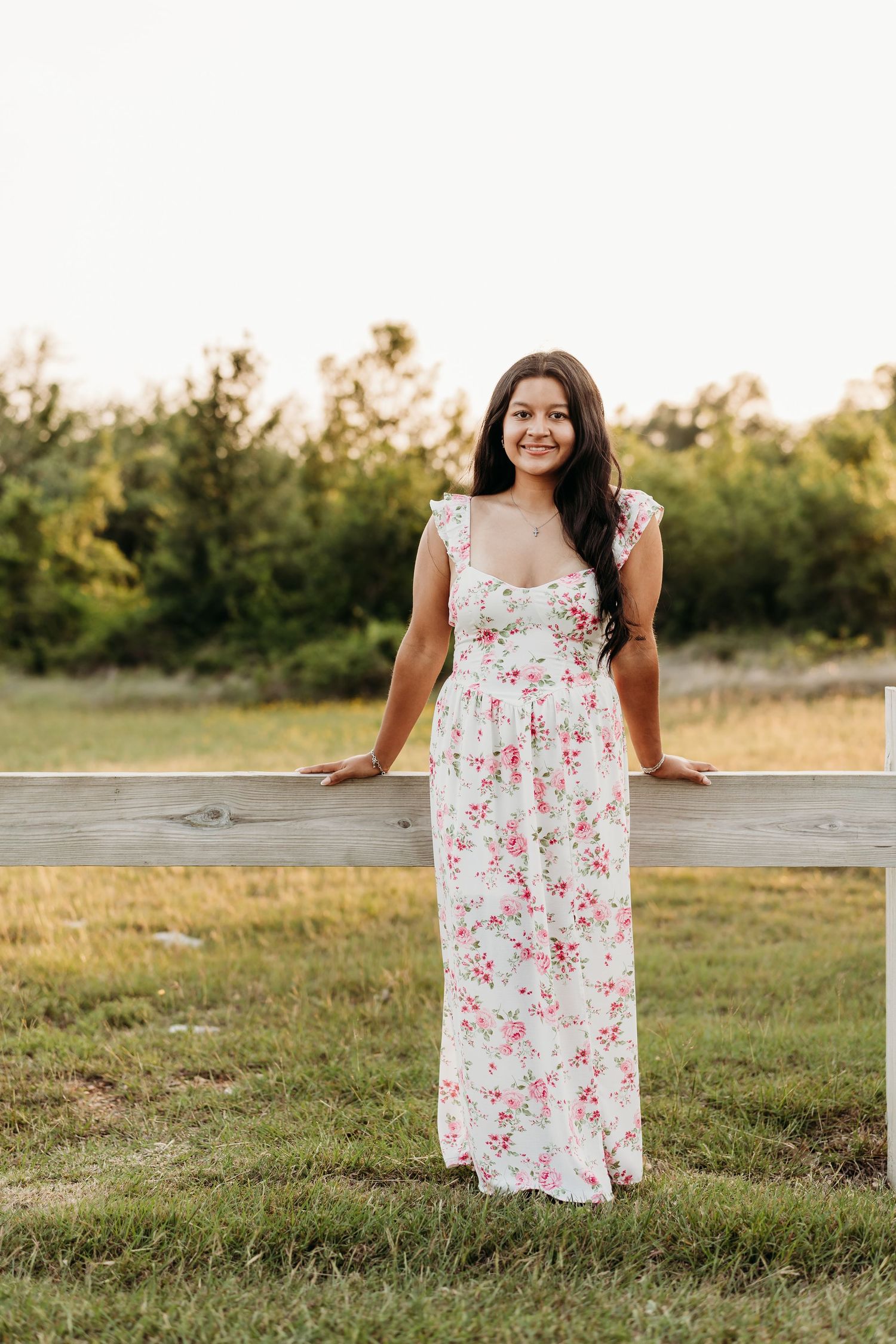 Young woman in white floral maxi dress smiling by a wooden fence in a sunny outdoor field.