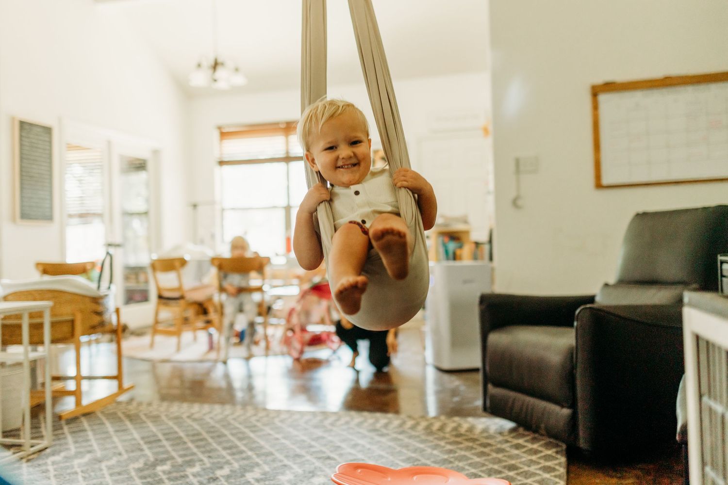 Happy toddler swinging in an indoor fabric baby swing in a bright living room.