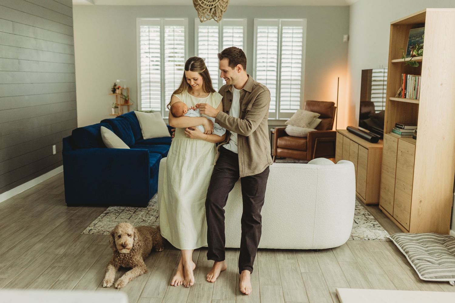 Parents standing barefoot in bright living room admiring their newborn baby with goldendoodle nearby.