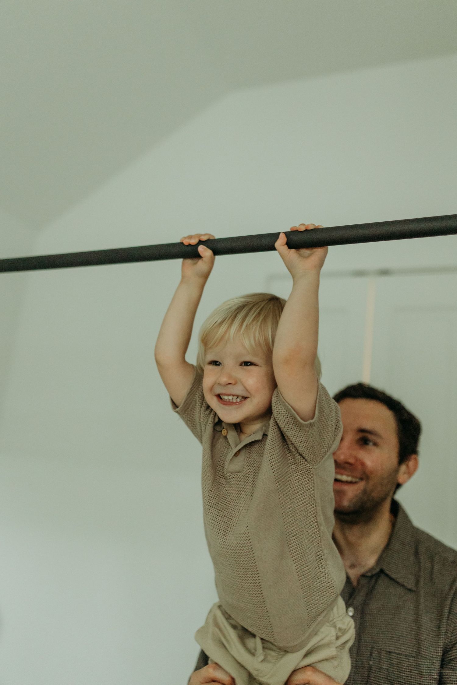Happy toddler hanging from a pull-up bar while smiling father assists during a playful indoor family moment.