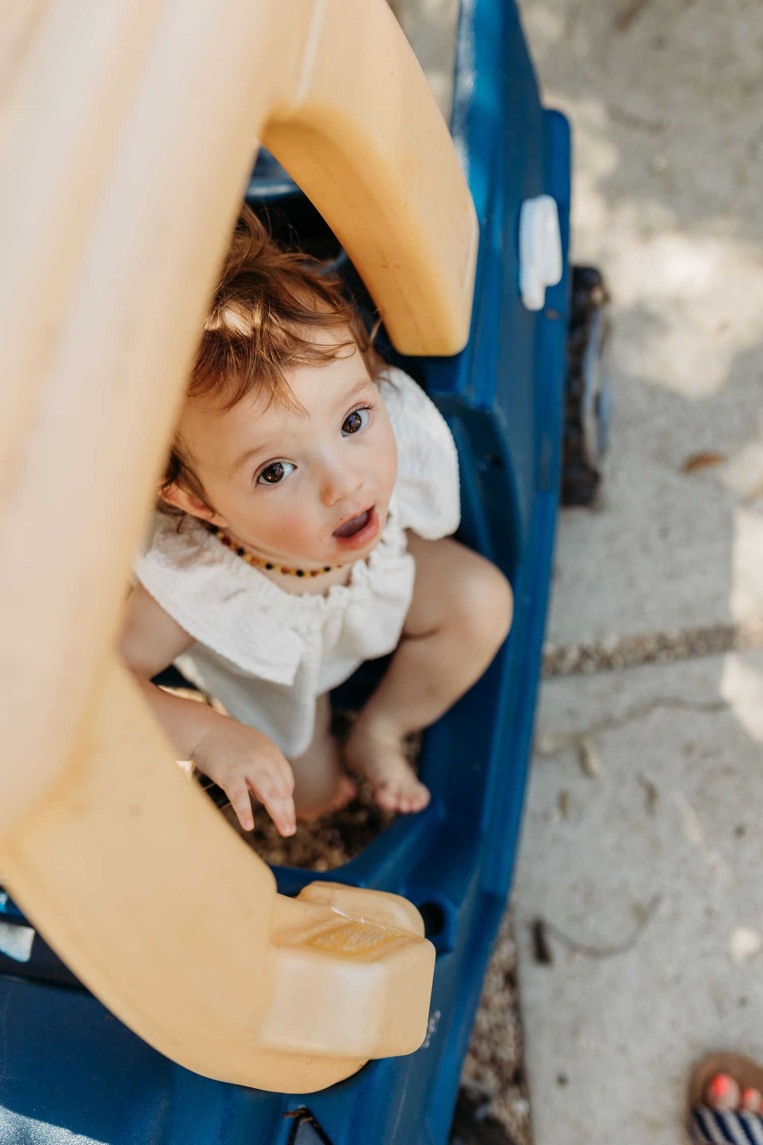 An adorable toddler in a white dress sits inside a blue plastic toy car looking up curiously.