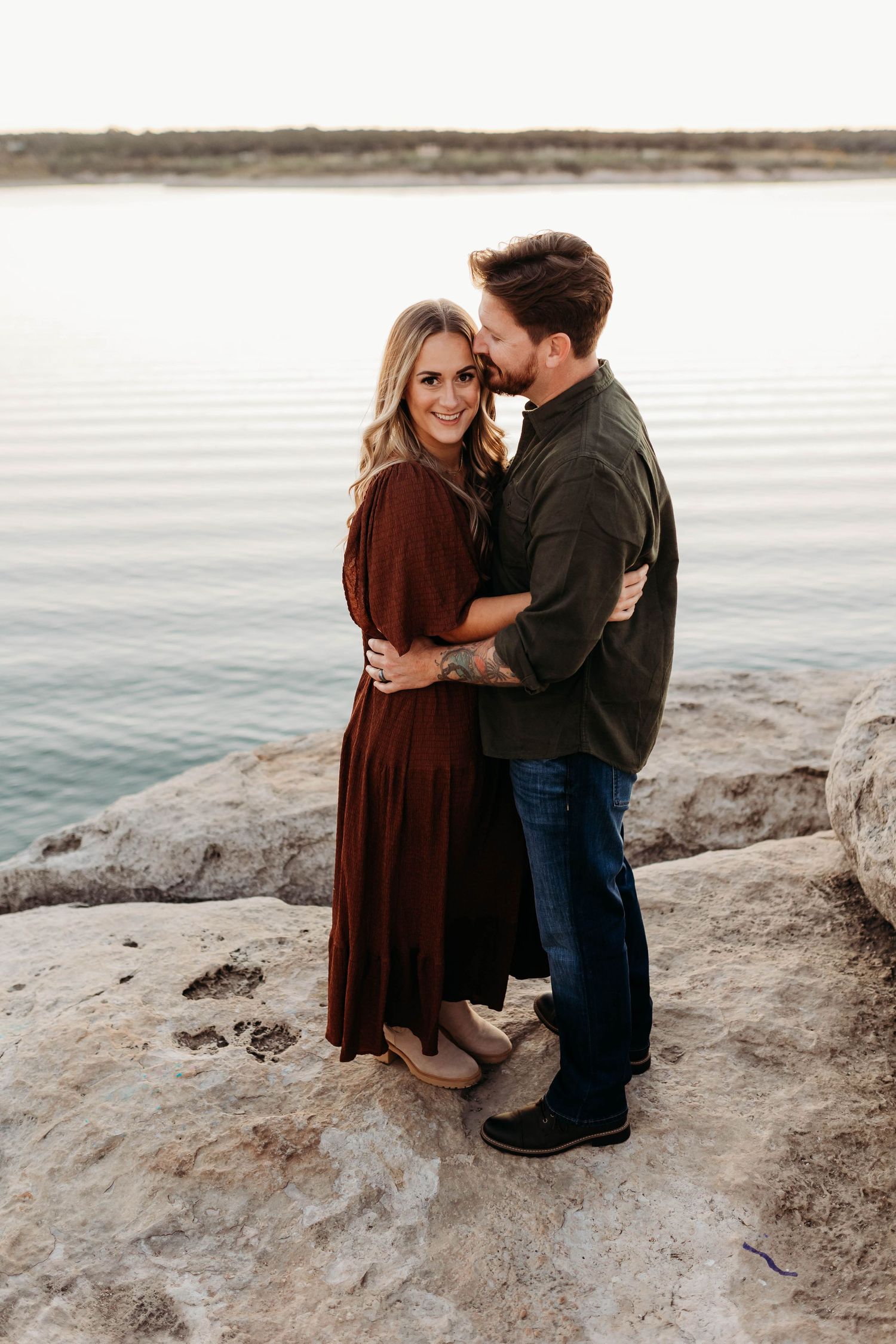Couple embracing on rocky lakeside, woman in burgundy dress smiling at camera during golden hour engagement photo.