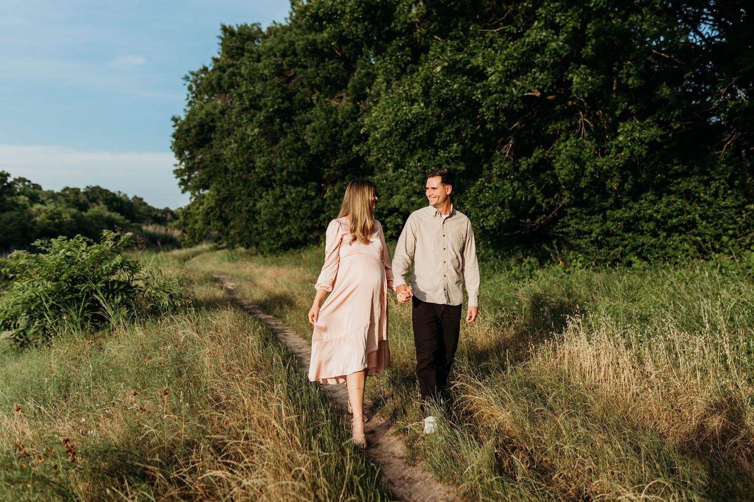 A couple walks hand in hand along a scenic path through lush green fields under a blue sky.