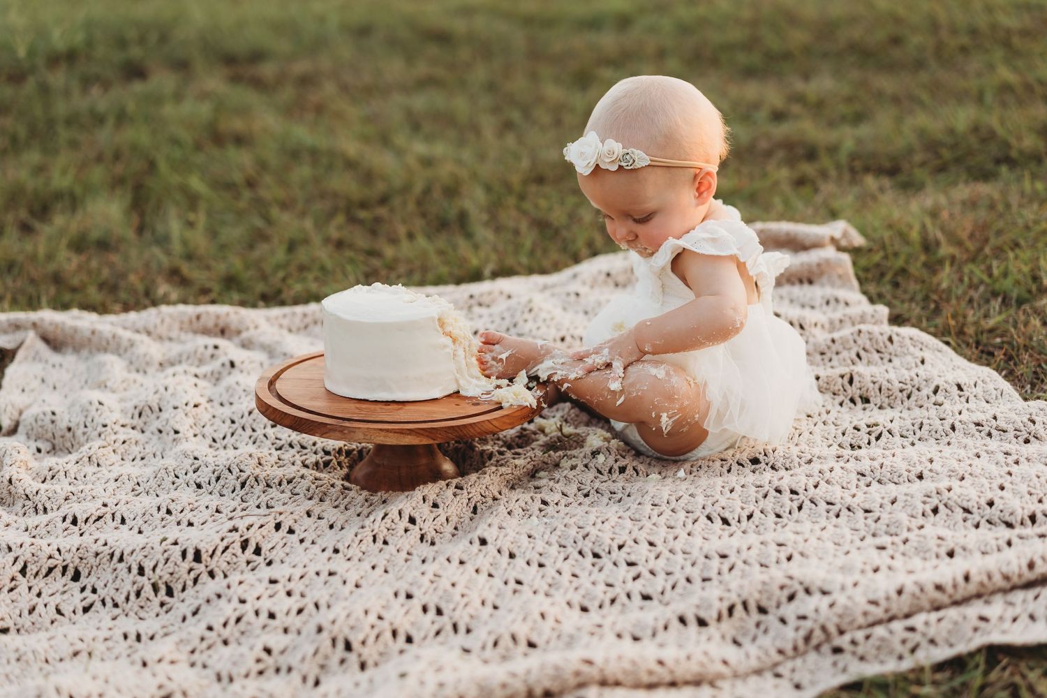 Baby girl in white dress and floral headband reaching for smash cake on blanket.