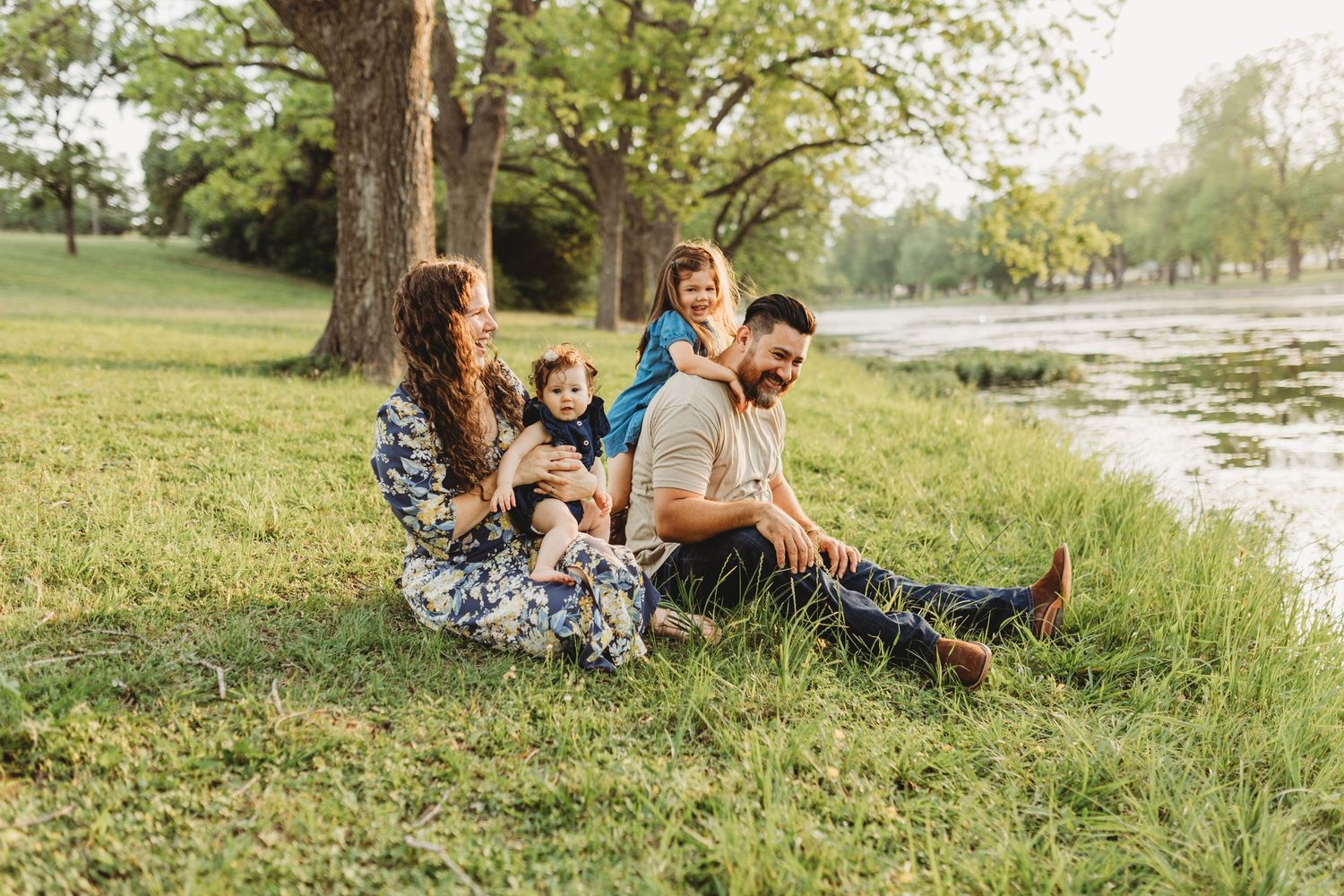 Happy family with two young children sitting together on green grass by a pond under large trees.