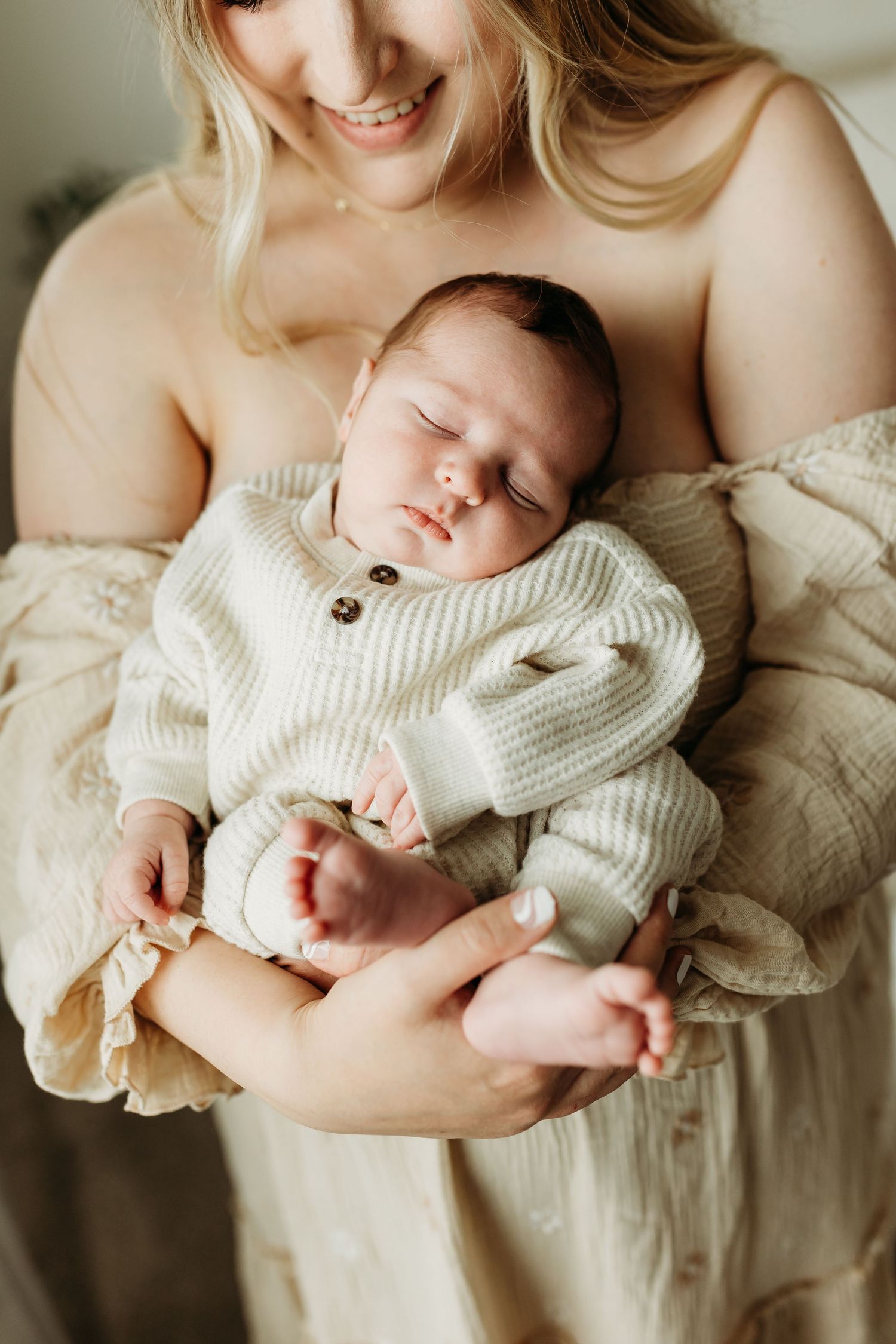 Smiling mother cradles sleeping newborn in cream knit onesie in her arms.