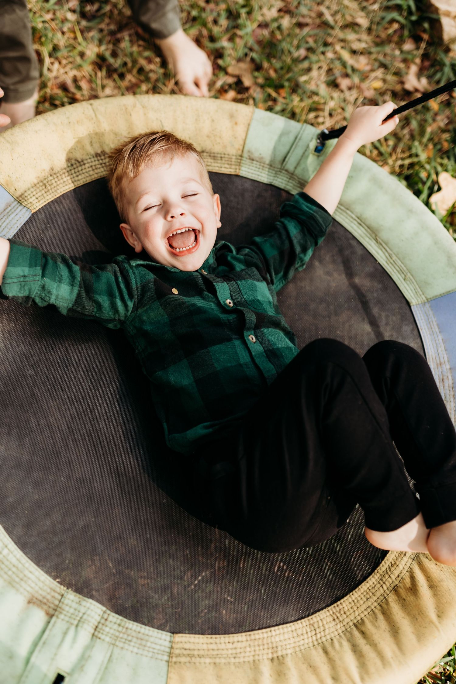 Laughing young boy in green plaid shirt lying on a small trampoline outdoors.