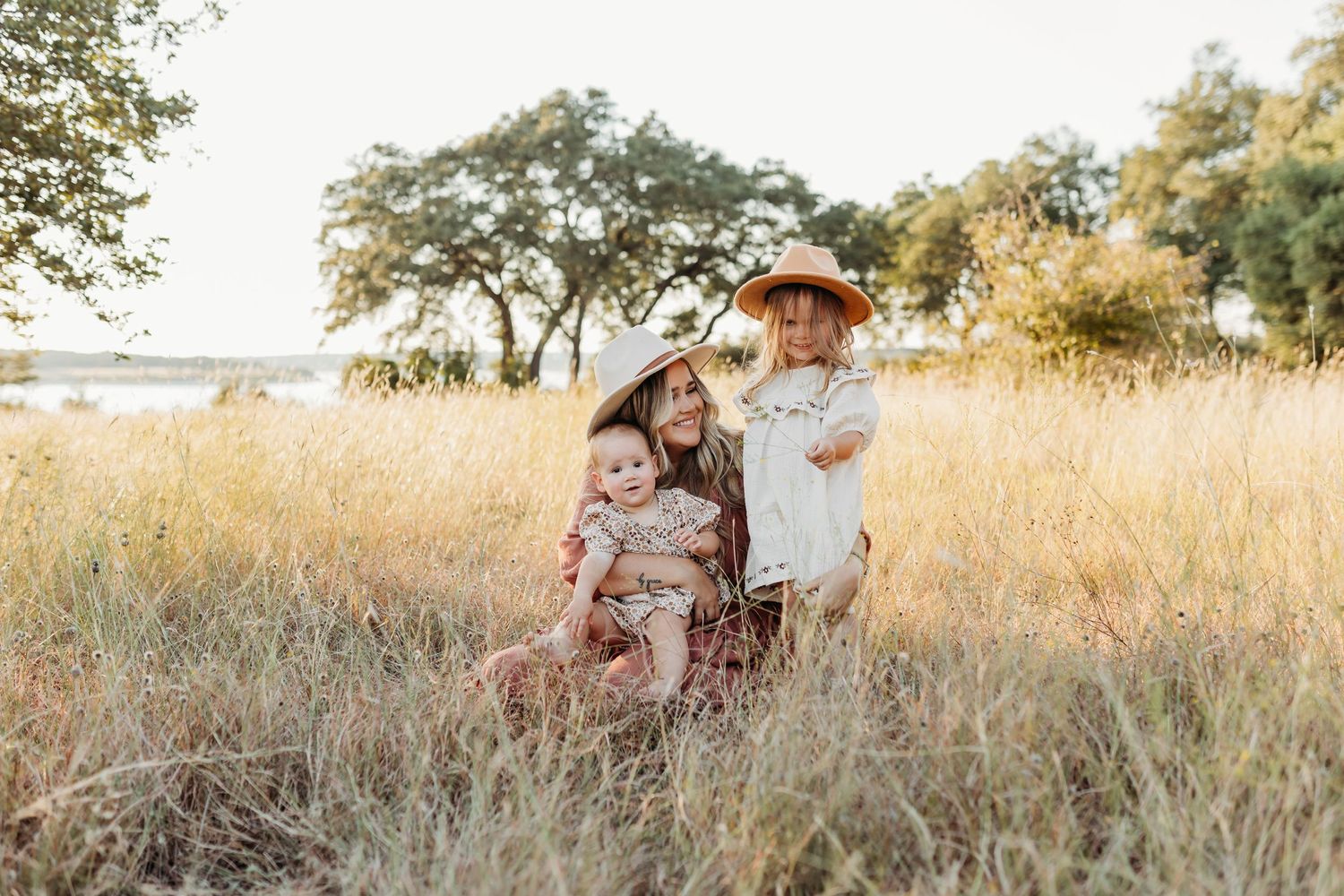 Mother sitting in golden grass field with three young children on sunny day.