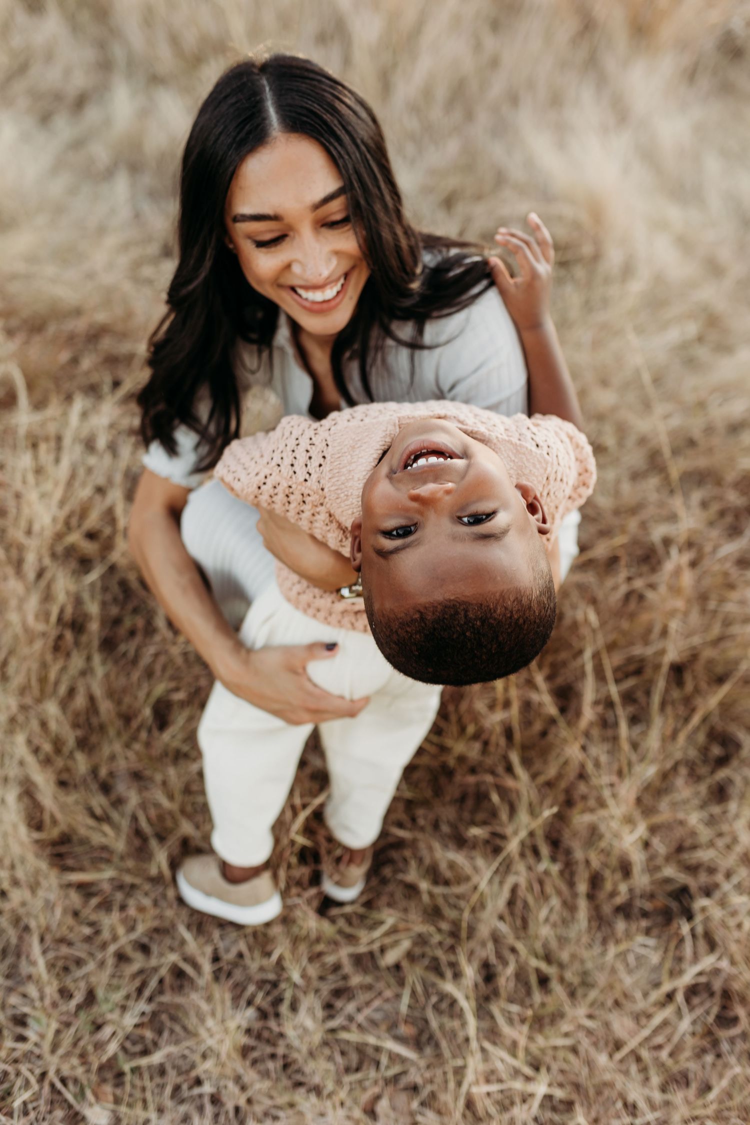 A smiling mother playfully holds her laughing toddler upside down in a dry golden grass field.