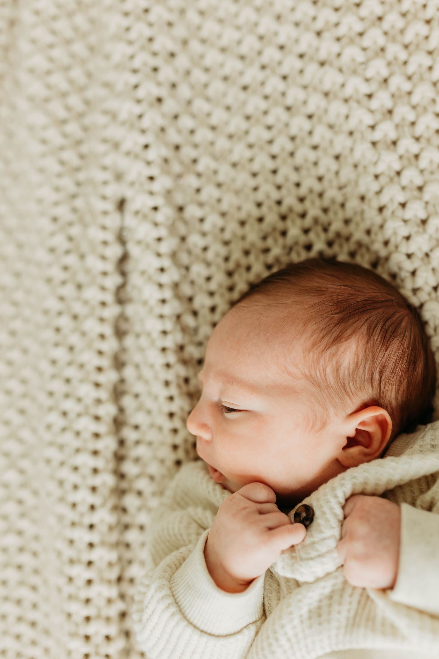 Peaceful newborn baby resting on a cream knit blanket in soft natural light.