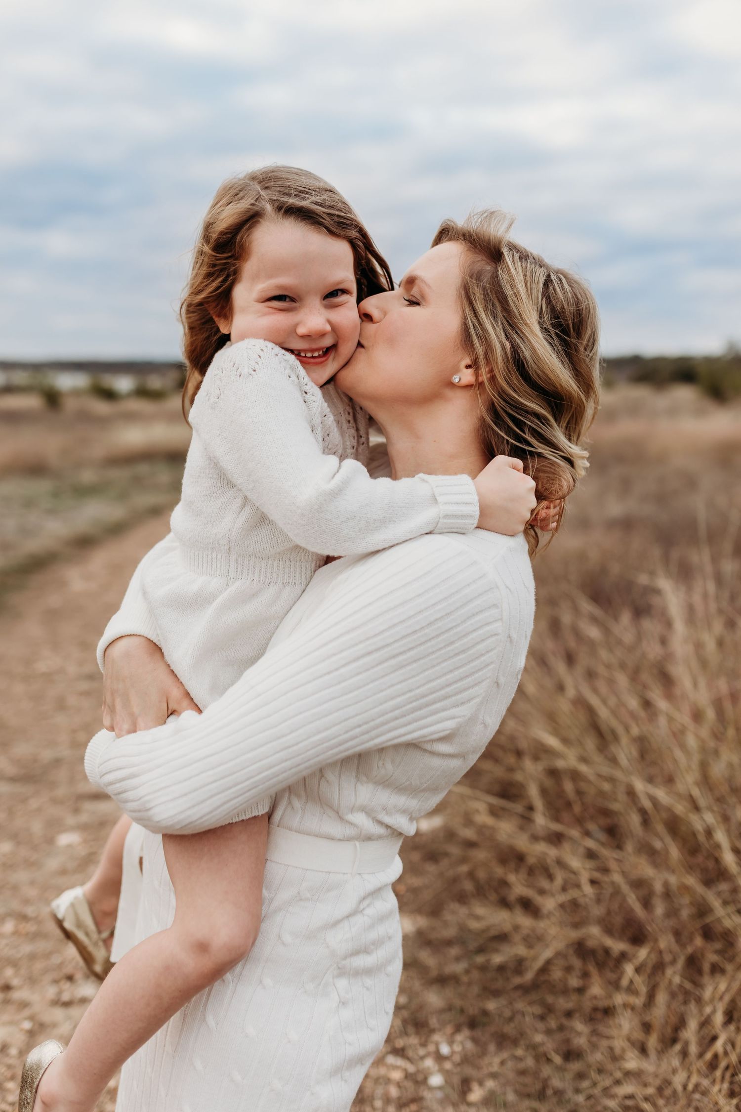 A mother kisses her smiling young daughter while holding her in a field.