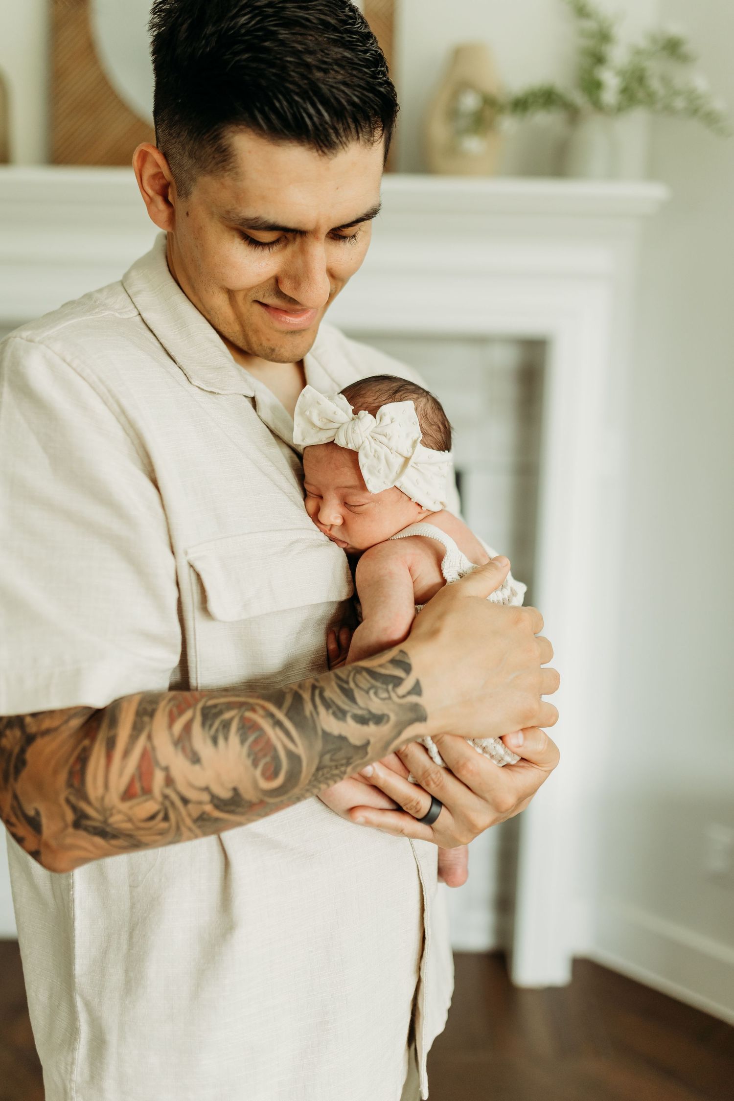 Tattooed father tenderly holding sleeping newborn baby girl with white bow headband against his chest.