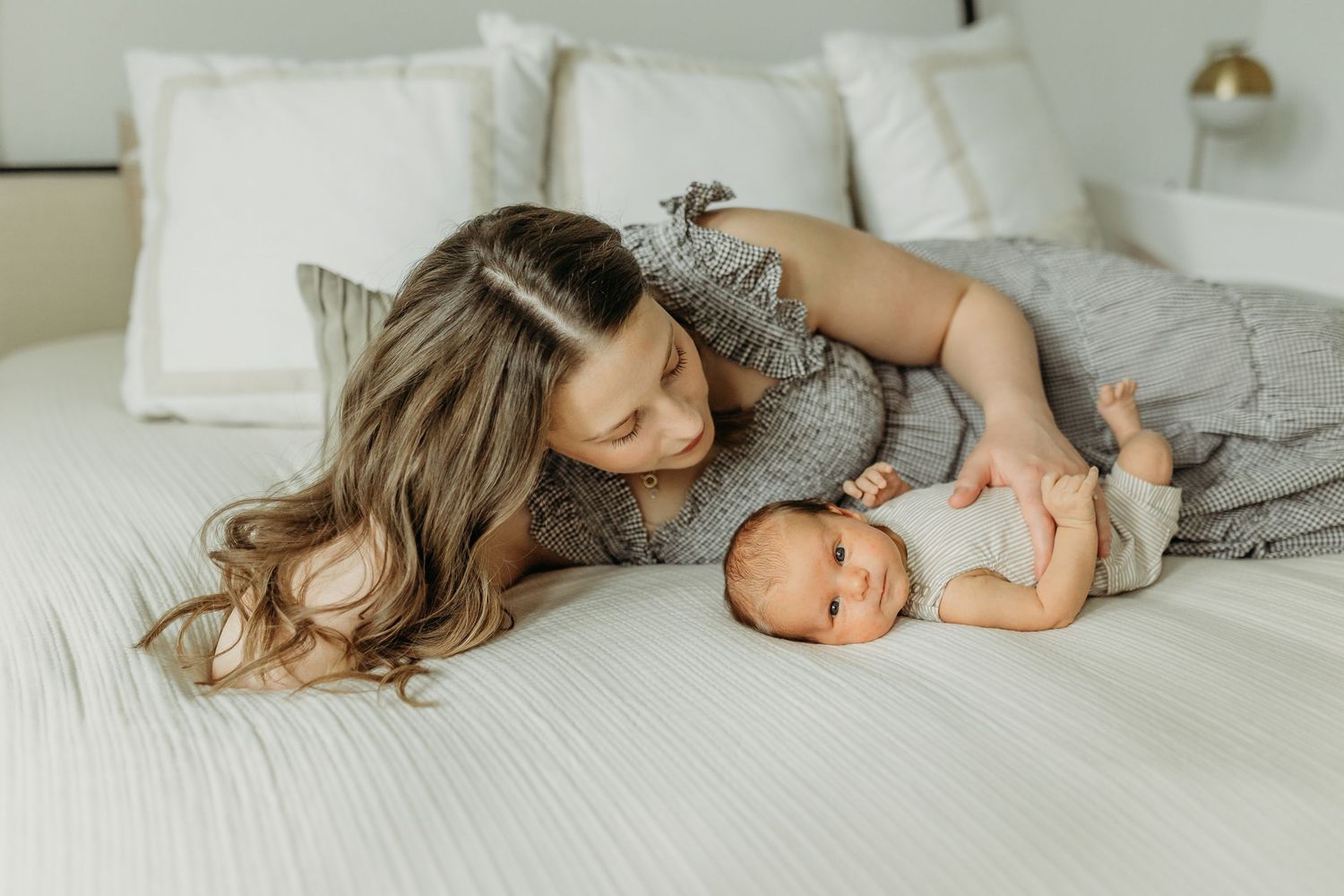 Mother lying on white bed smiling at her newborn baby during a tender indoor lifestyle photography session.