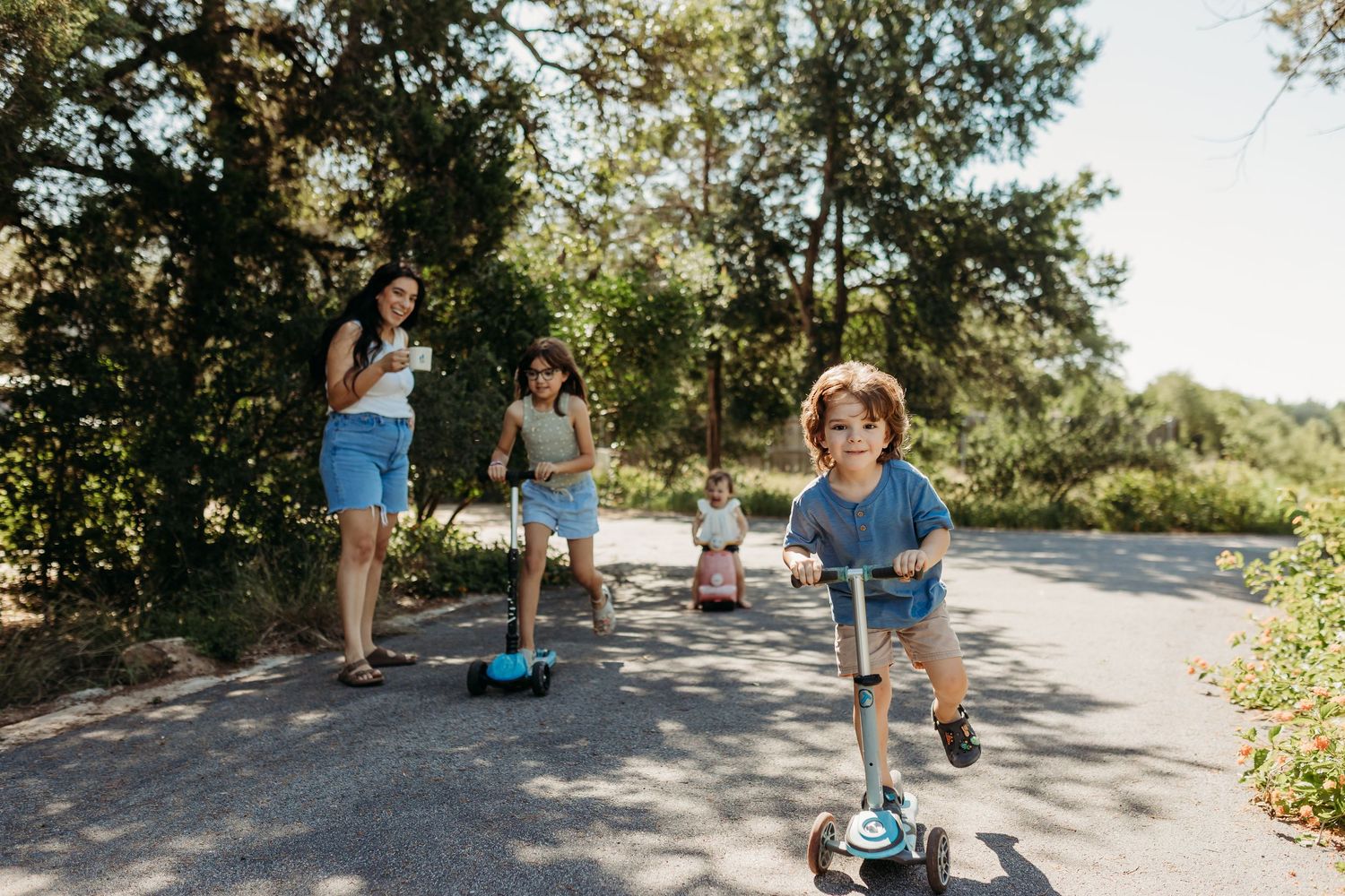 A mother watches her children ride scooters along a sunny tree-lined path outdoors.