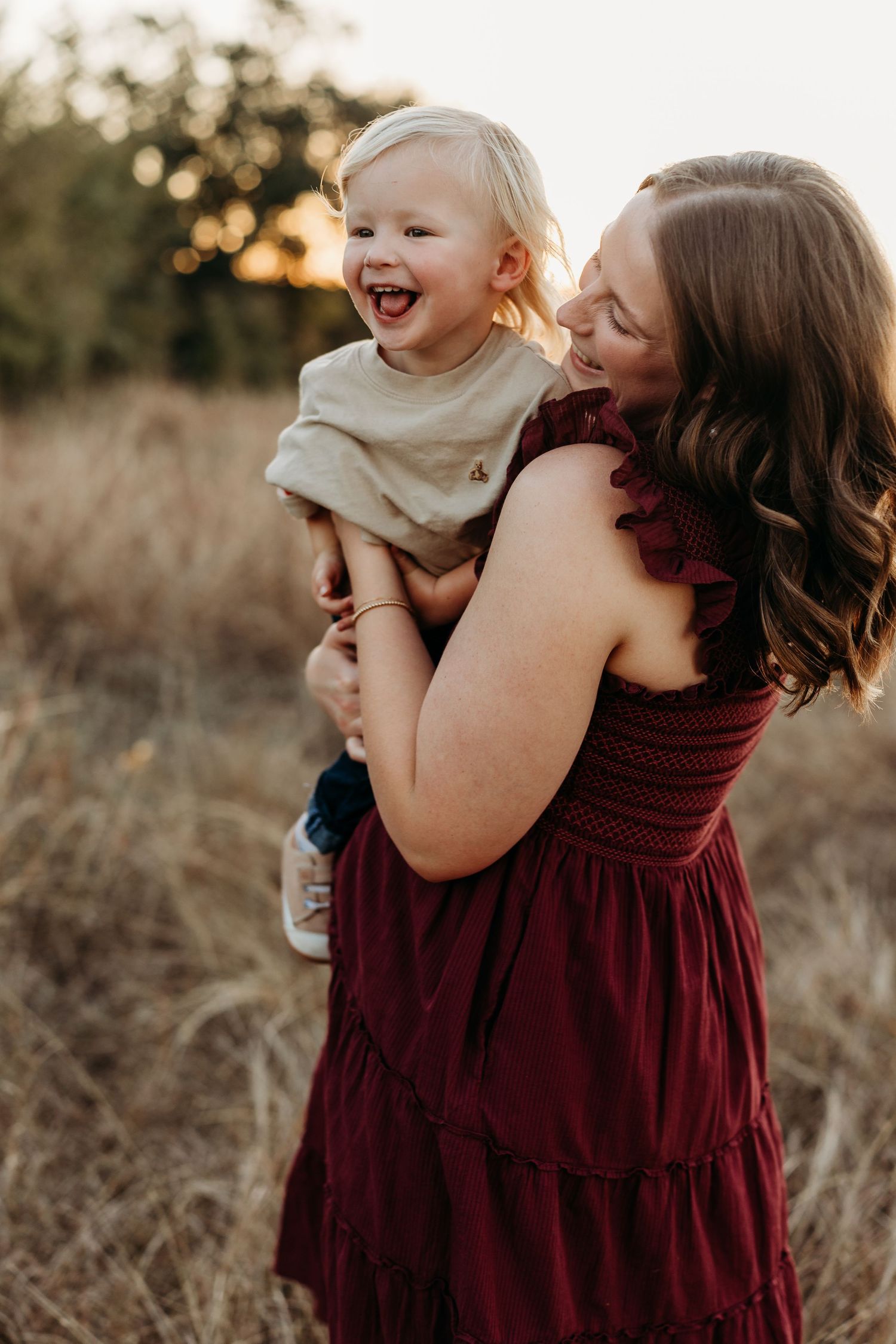 Joyful mother in burgundy dress holding her laughing blonde toddler in a sunlit field.