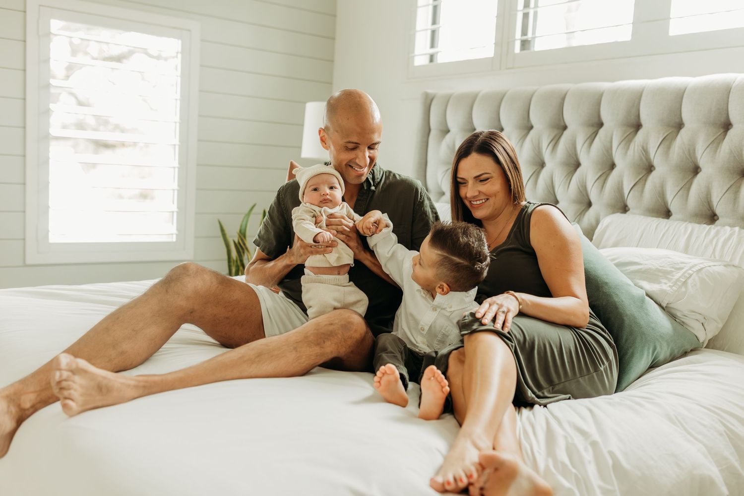 Happy family of four relaxes together on white bed in bright modern bedroom during photo session.