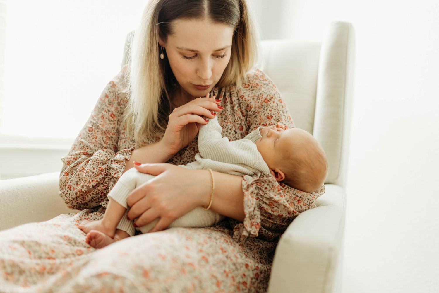 Blonde mother in floral dress tenderly holding and gazing at sleeping newborn baby.