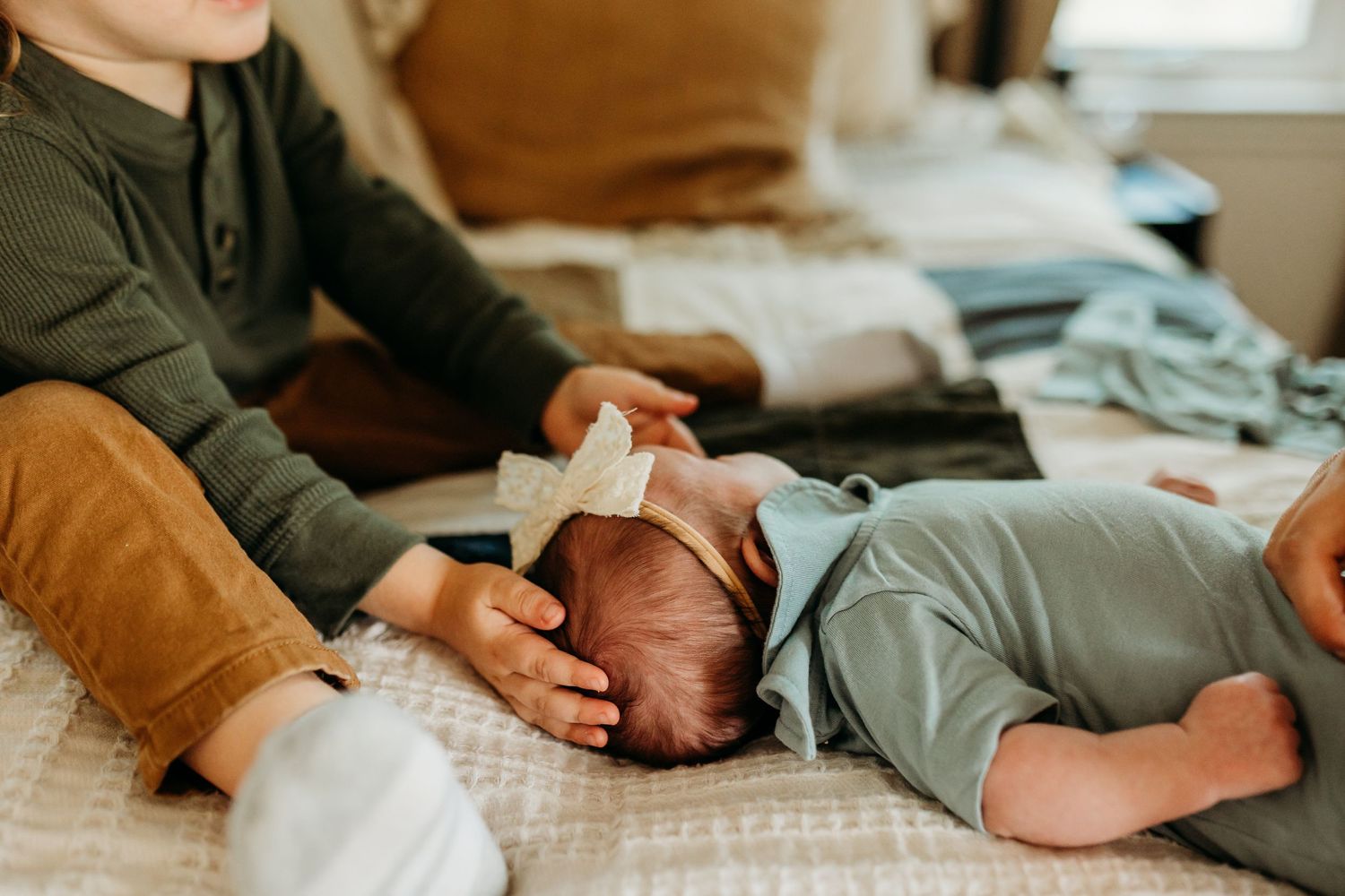 An older sibling tenderly touches a newborn baby wearing a white bow headband while sitting on a bed together.