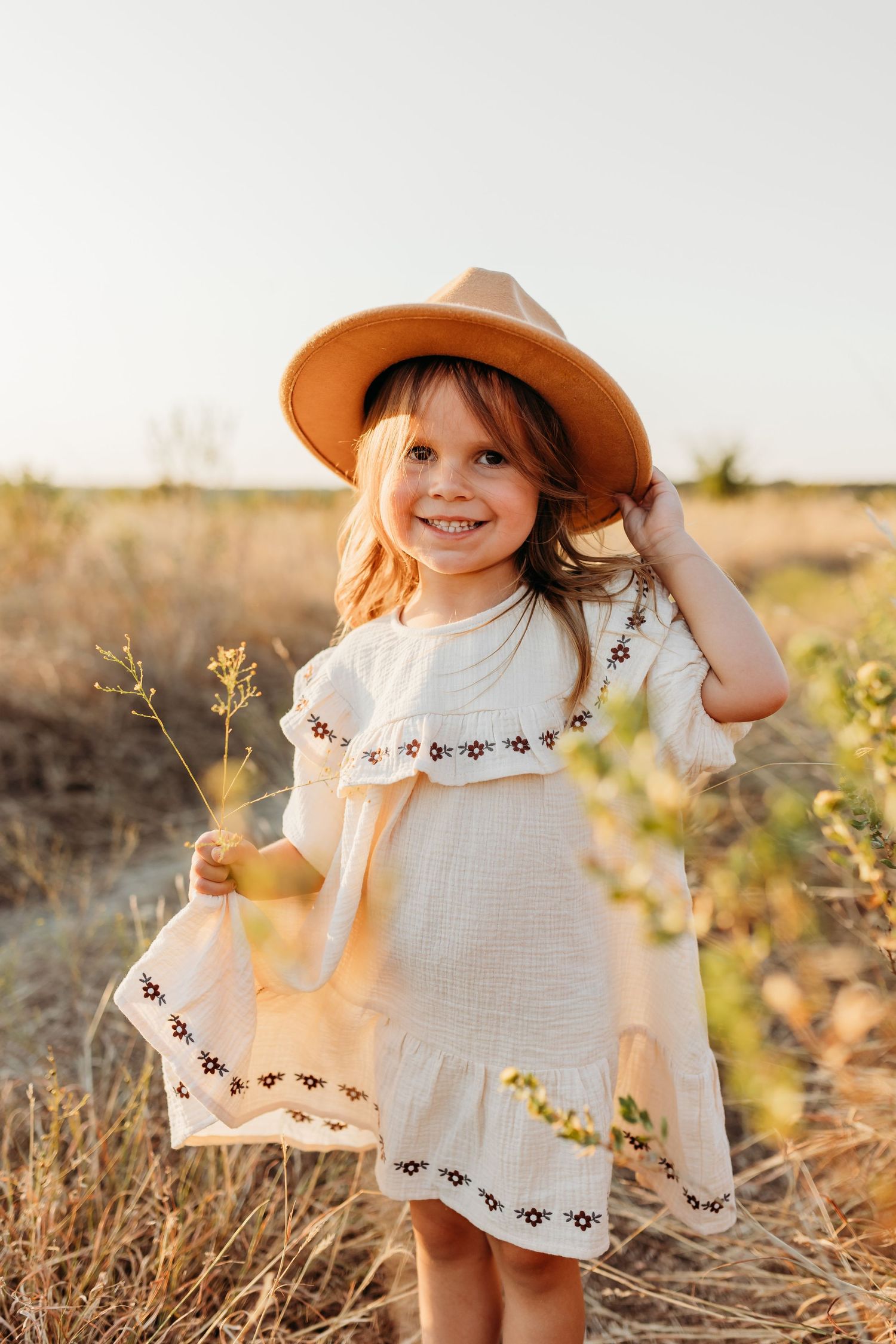 Smiling little girl in white embroidered dress and tan hat holding wildflowers in a sunny field.