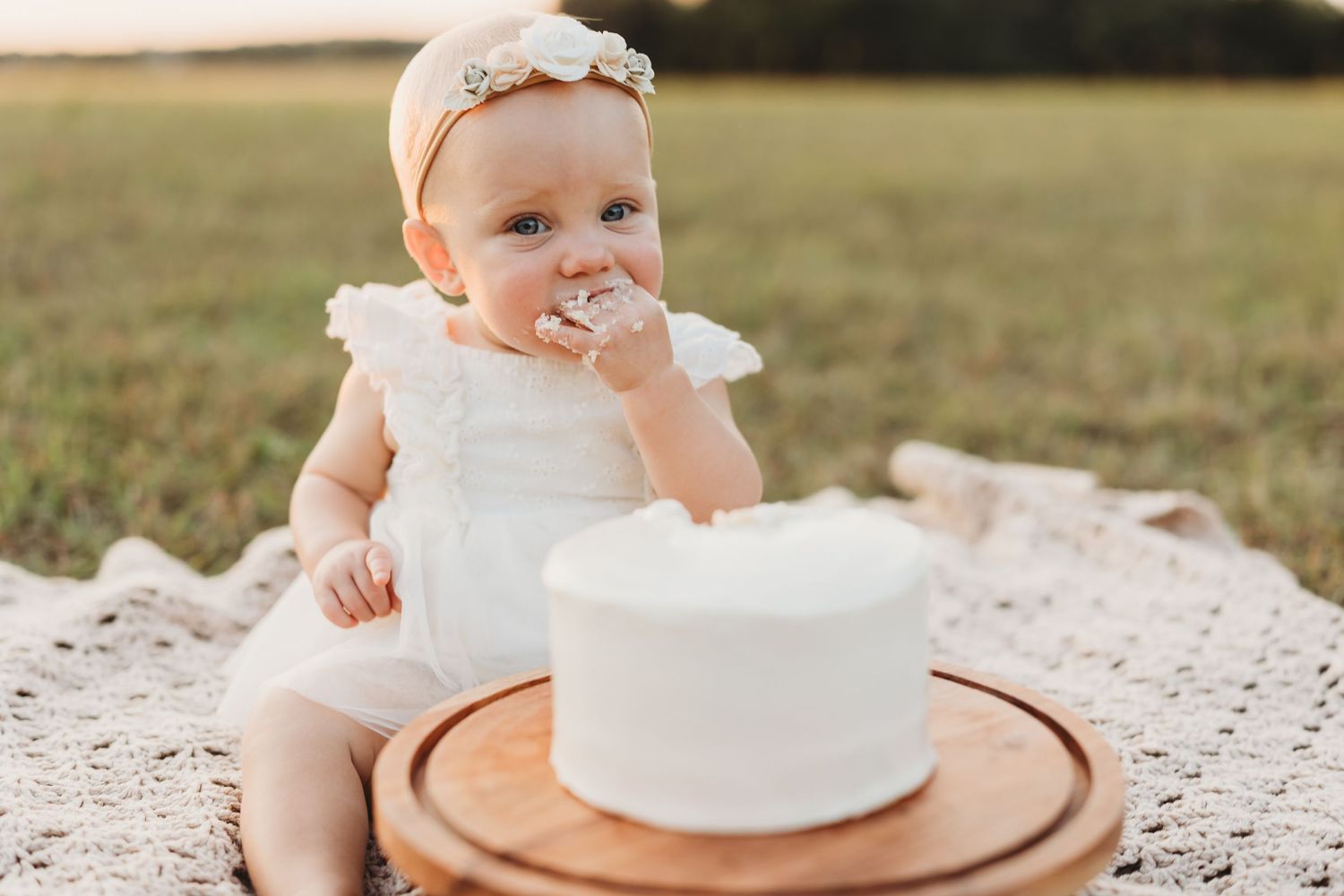 Baby girl in white dress eating first birthday smash cake outdoors in a field.
