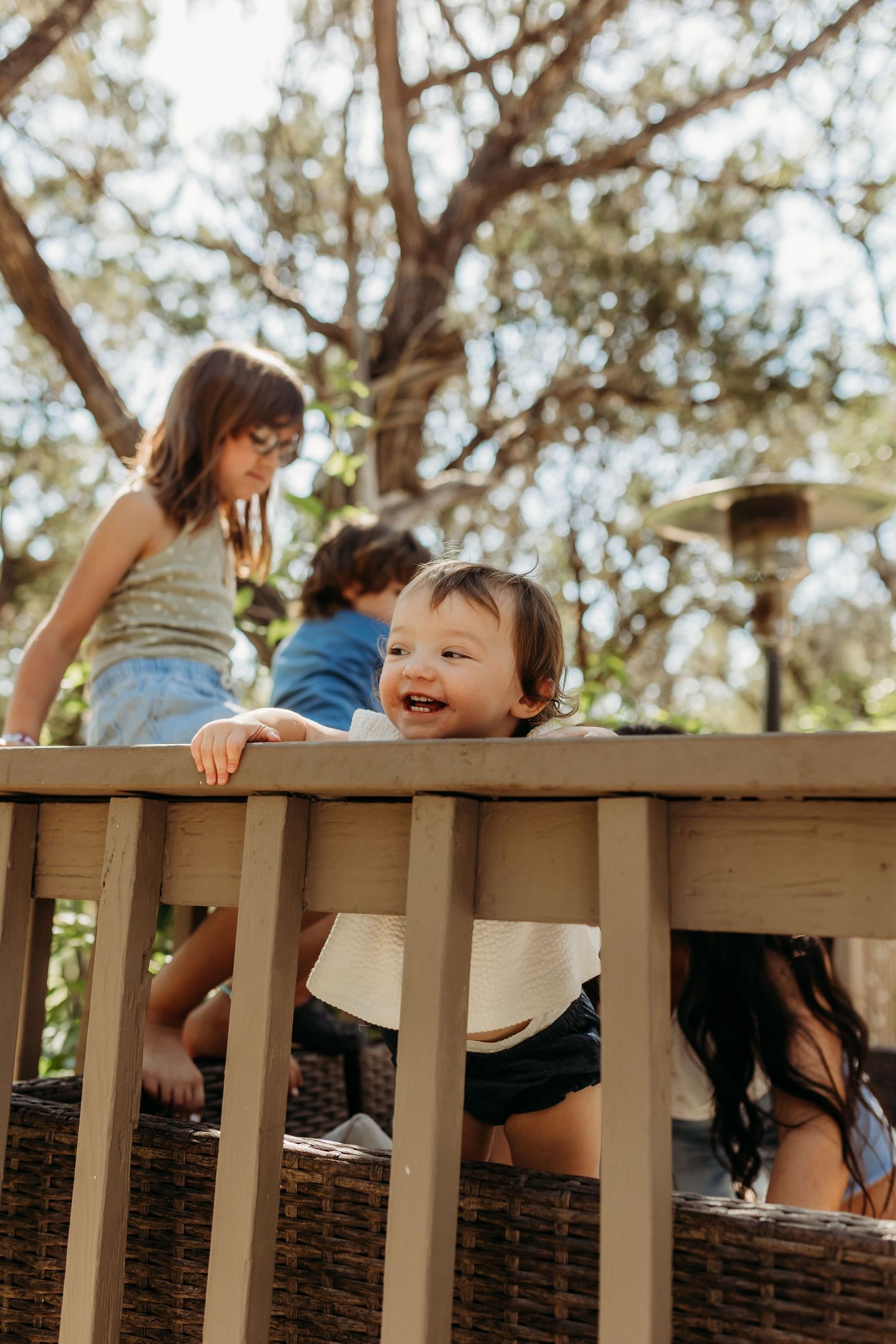 A happy toddler peeks through wooden deck railings while siblings play on a sunny outdoor patio.