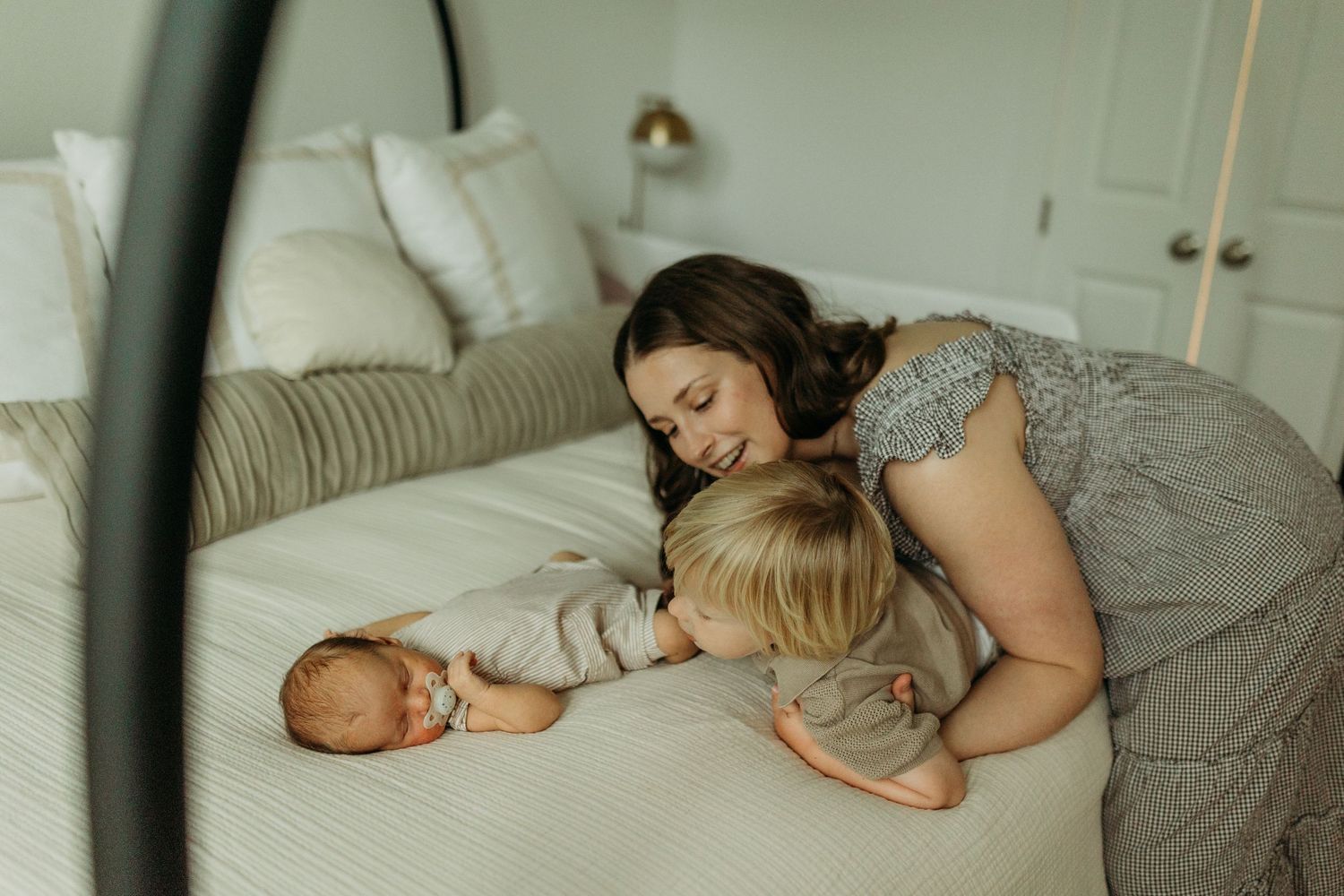 Mother and toddler leaning over a sleeping newborn on a white bed during a cozy newborn session.