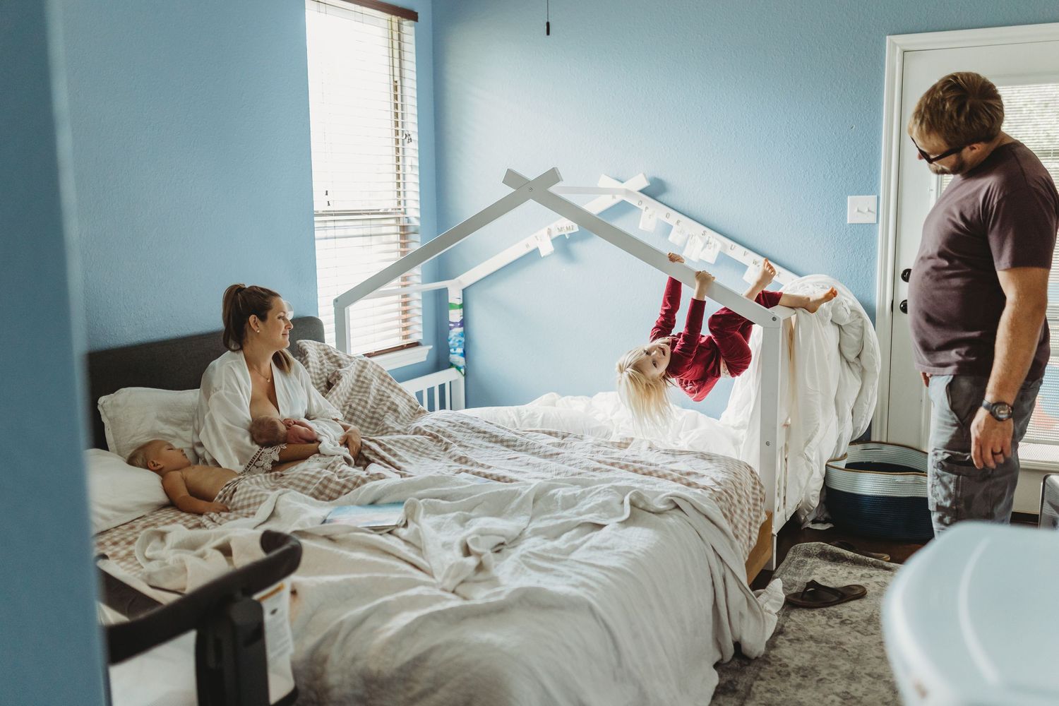 Family with toddler playfully hanging off bed beside mother and newborn in hospital room.