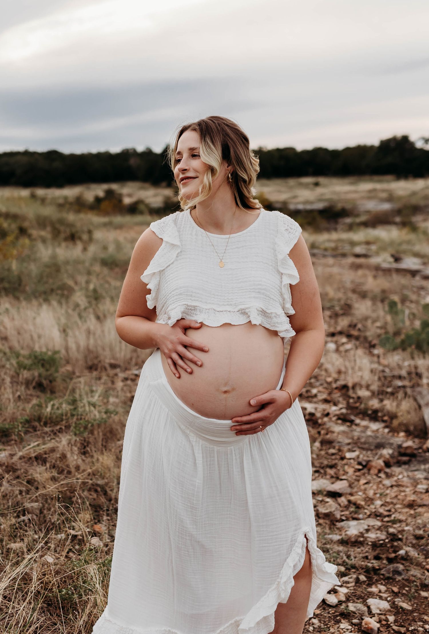 Pregnant woman in white crop top and skirt cradling her bare baby bump in an outdoor field setting.