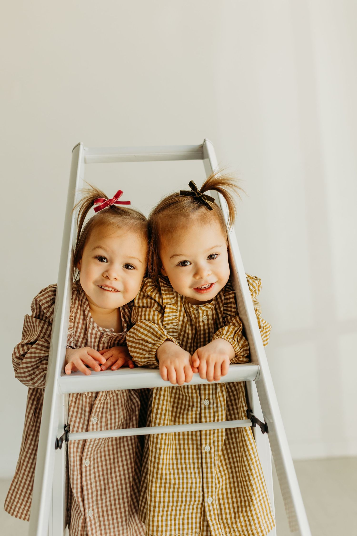 Two smiling toddler girls in matching plaid dresses lean on white ladder in bright white studio.