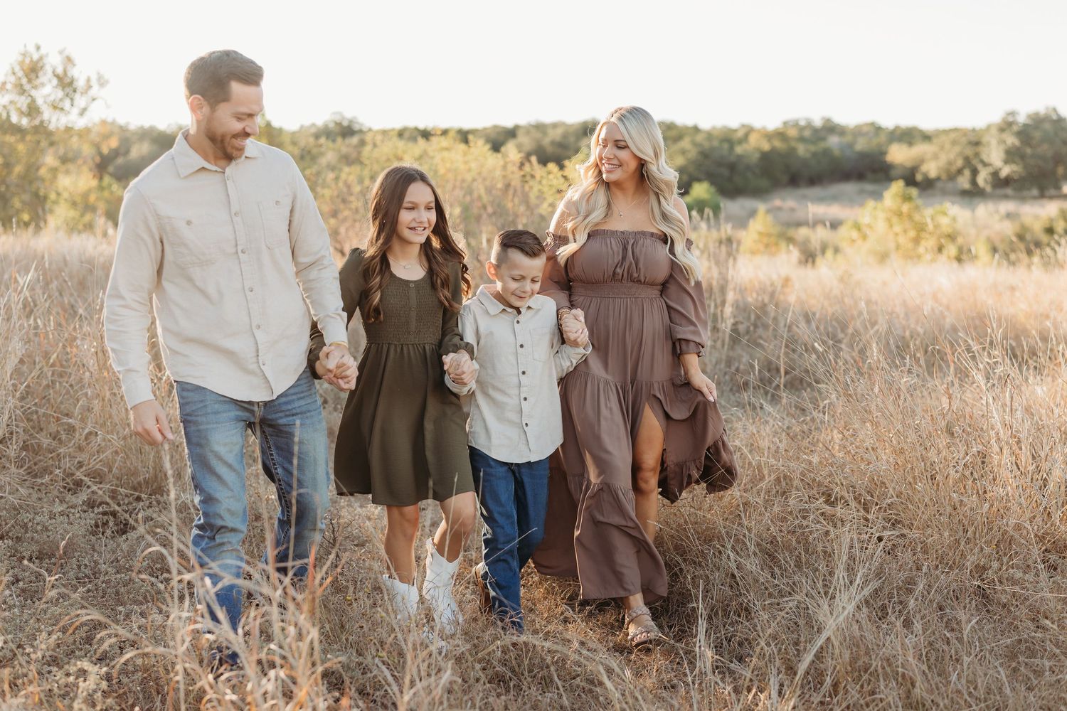 Family of four walking hand-in-hand through a golden field during a warm sunset.