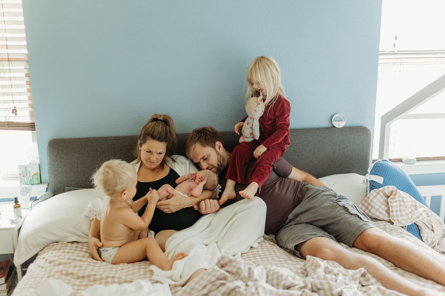 Happy family with newborn and two toddlers cuddling together on a bed with blue walls.