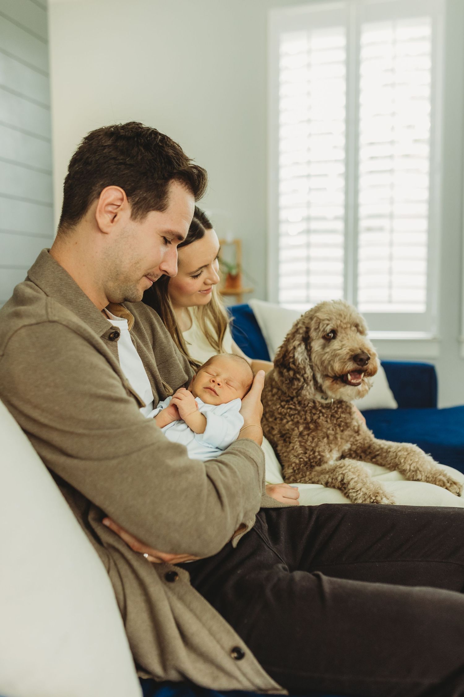 Family with newborn baby and curly brown dog relaxing together on white sofa at home.