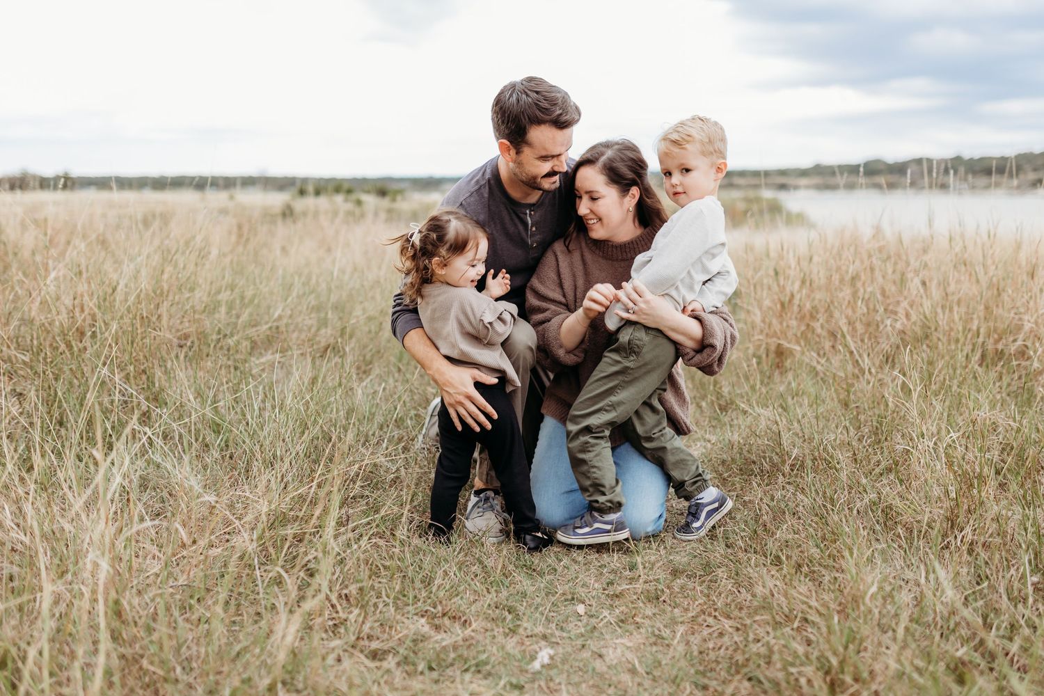 A loving family of four cuddles closely together in a tall grass field near a calm lake.