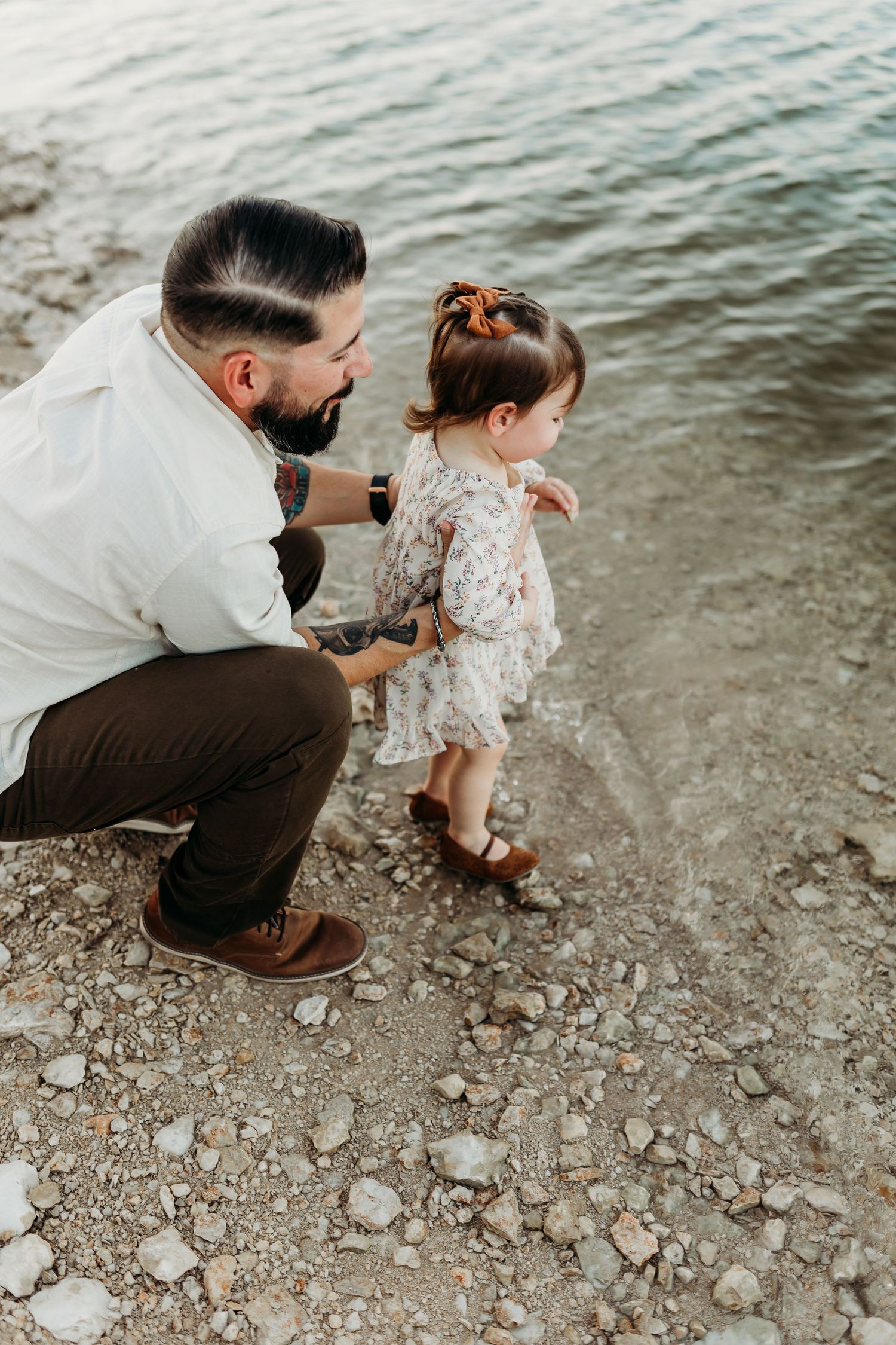 Father crouching beside toddler daughter at rocky waterside during family photo session.