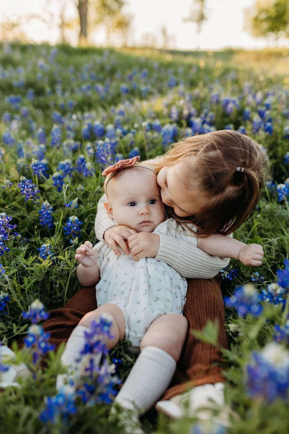 An older sister kisses a baby sibling while sitting in a field of bluebonnets.