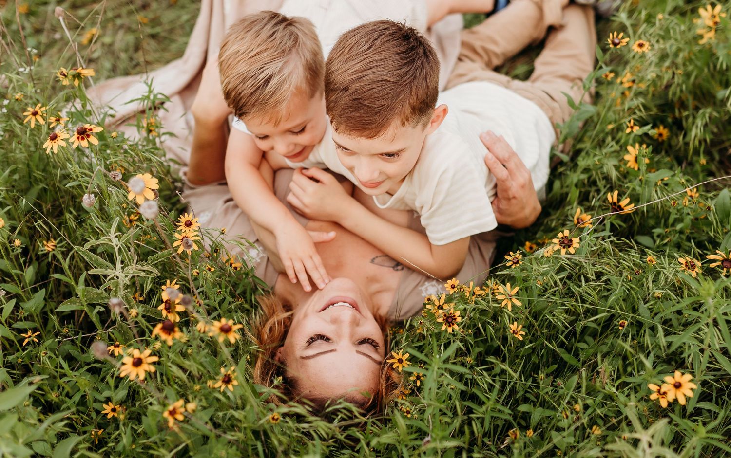 Two boys playfully cuddle with their laughing mother lying in a flower-filled meadow.