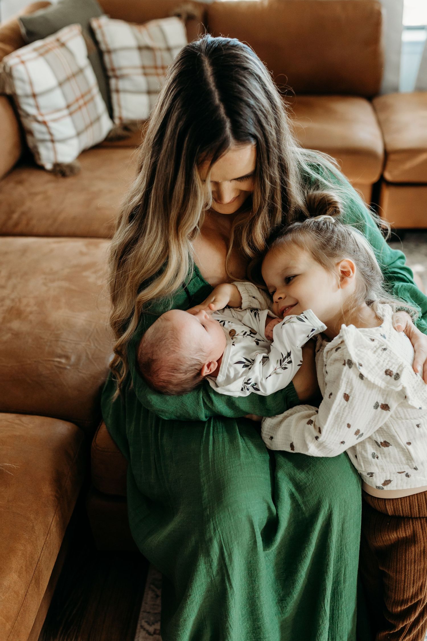 Mother in green dress tenderly holding newborn while toddler daughter leans in for a kiss indoors.