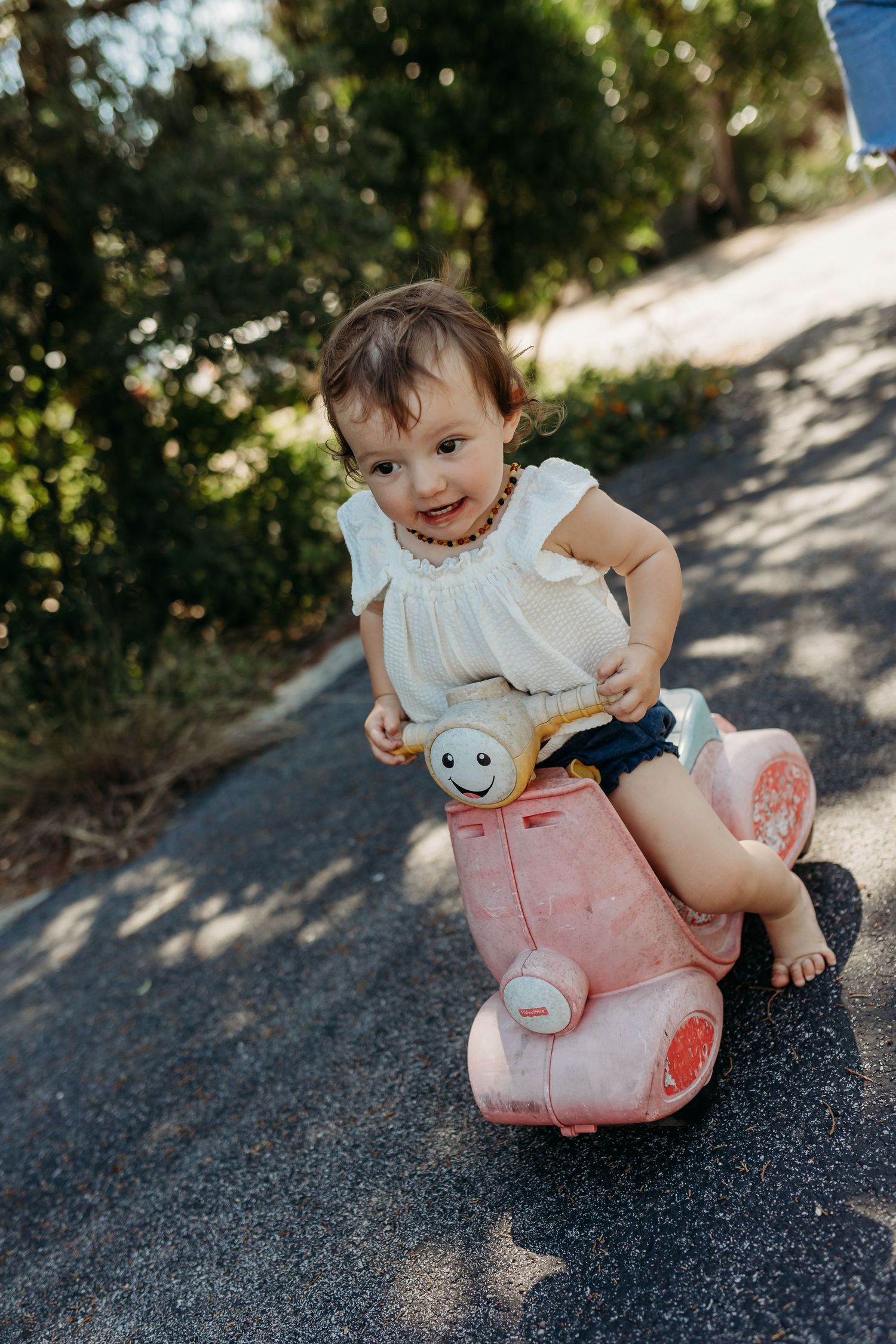 A cheerful toddler in a white dress rides a pink toy scooter on a sunlit outdoor path.