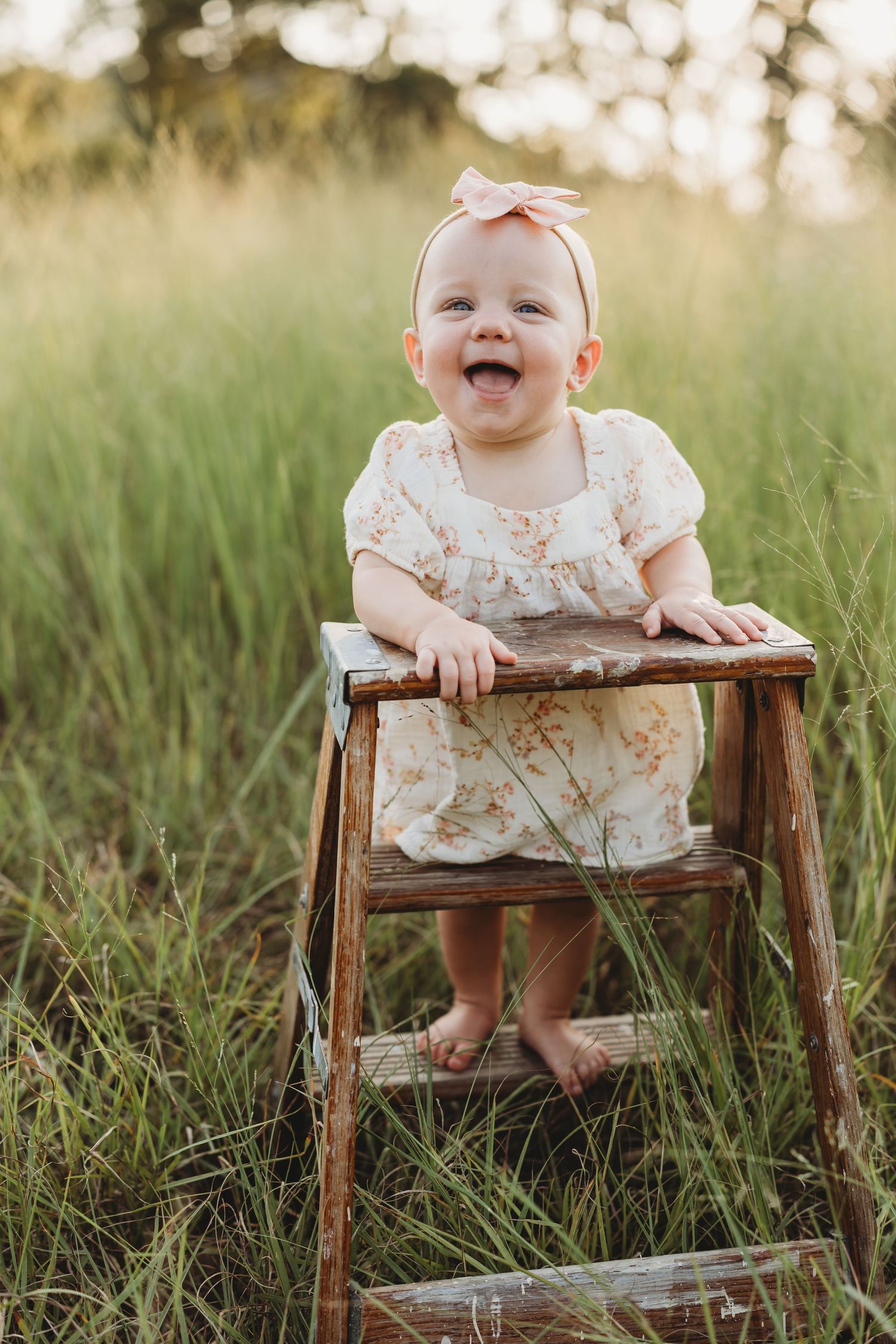 Laughing baby girl in floral romper standing on rustic wooden ladder in tall grass.