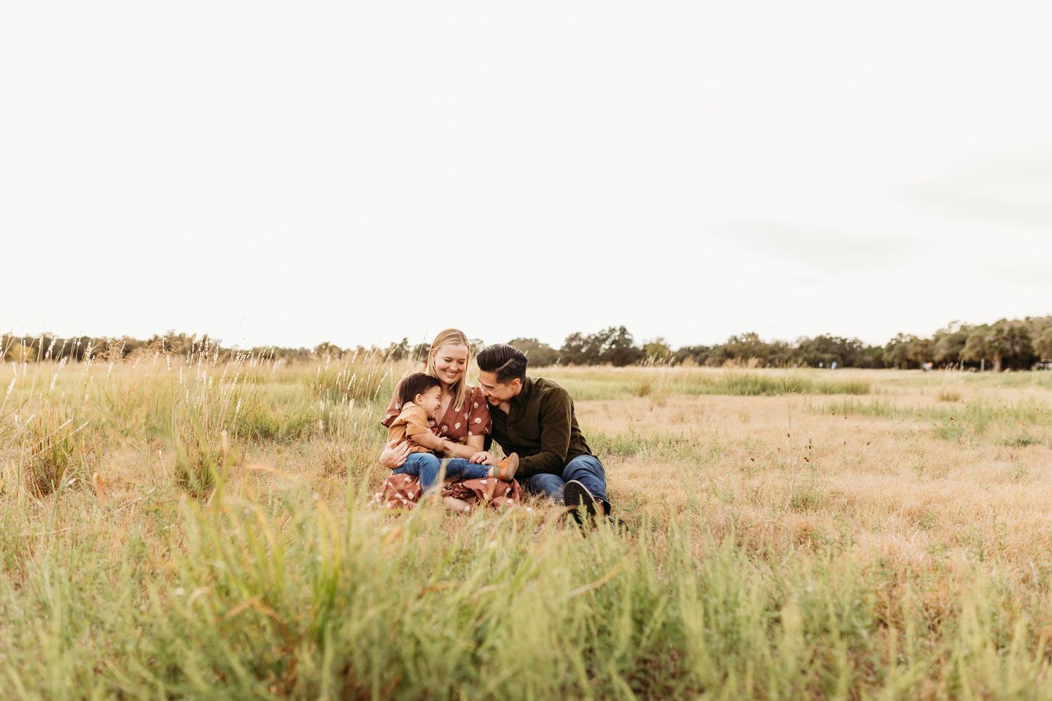 A loving family of three sits together in a lush green meadow sharing a warm, joyful moment outdoors.