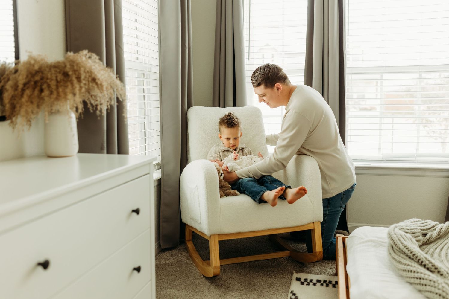 Father helps toddler sit in a white nursery rocking chair by a bright window.