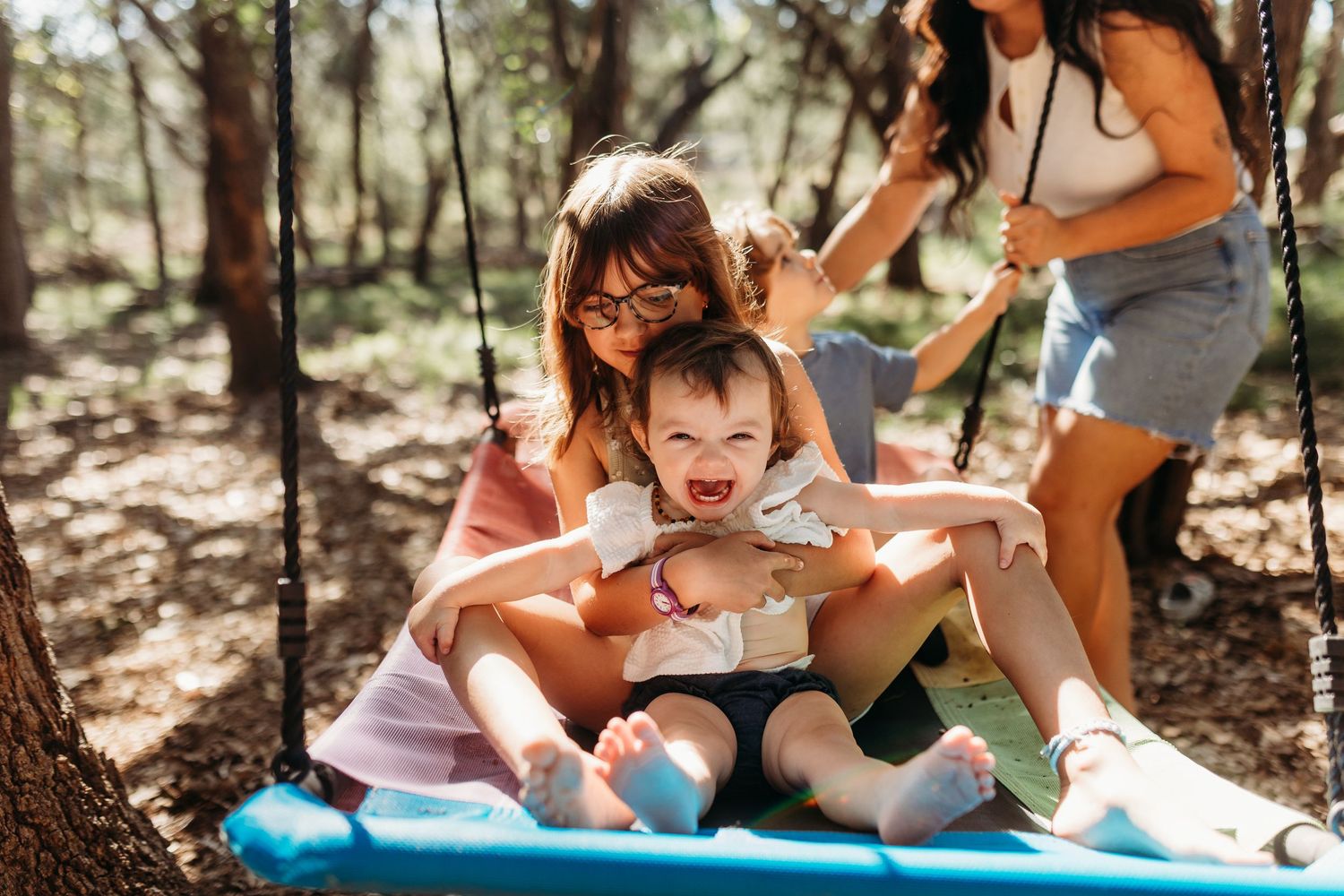 Woman and child laughing together on a large swing in a sunny woodland setting.