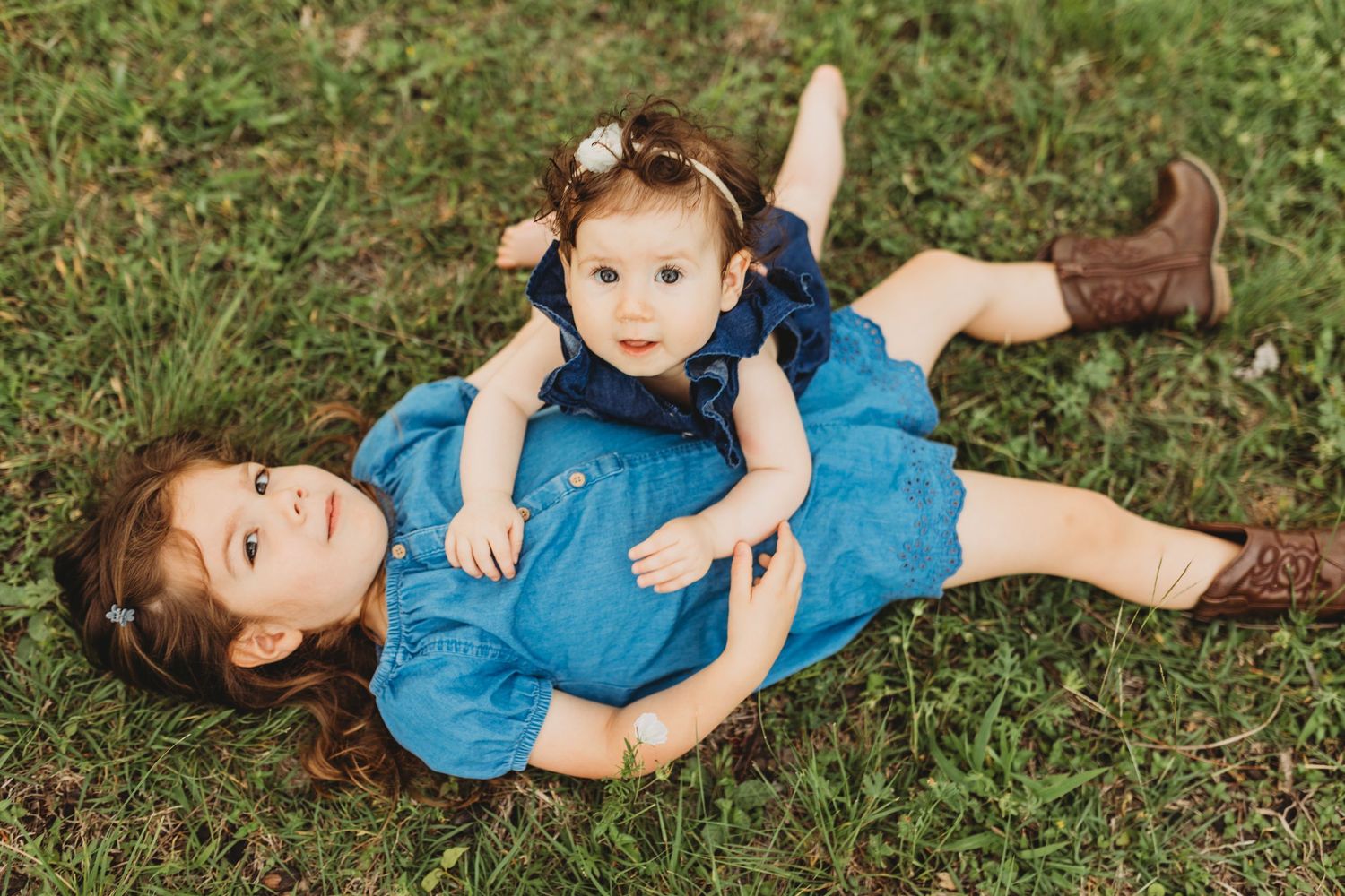 Two young sisters in blue denim dresses lying on green grass, older girl holding baby sister.