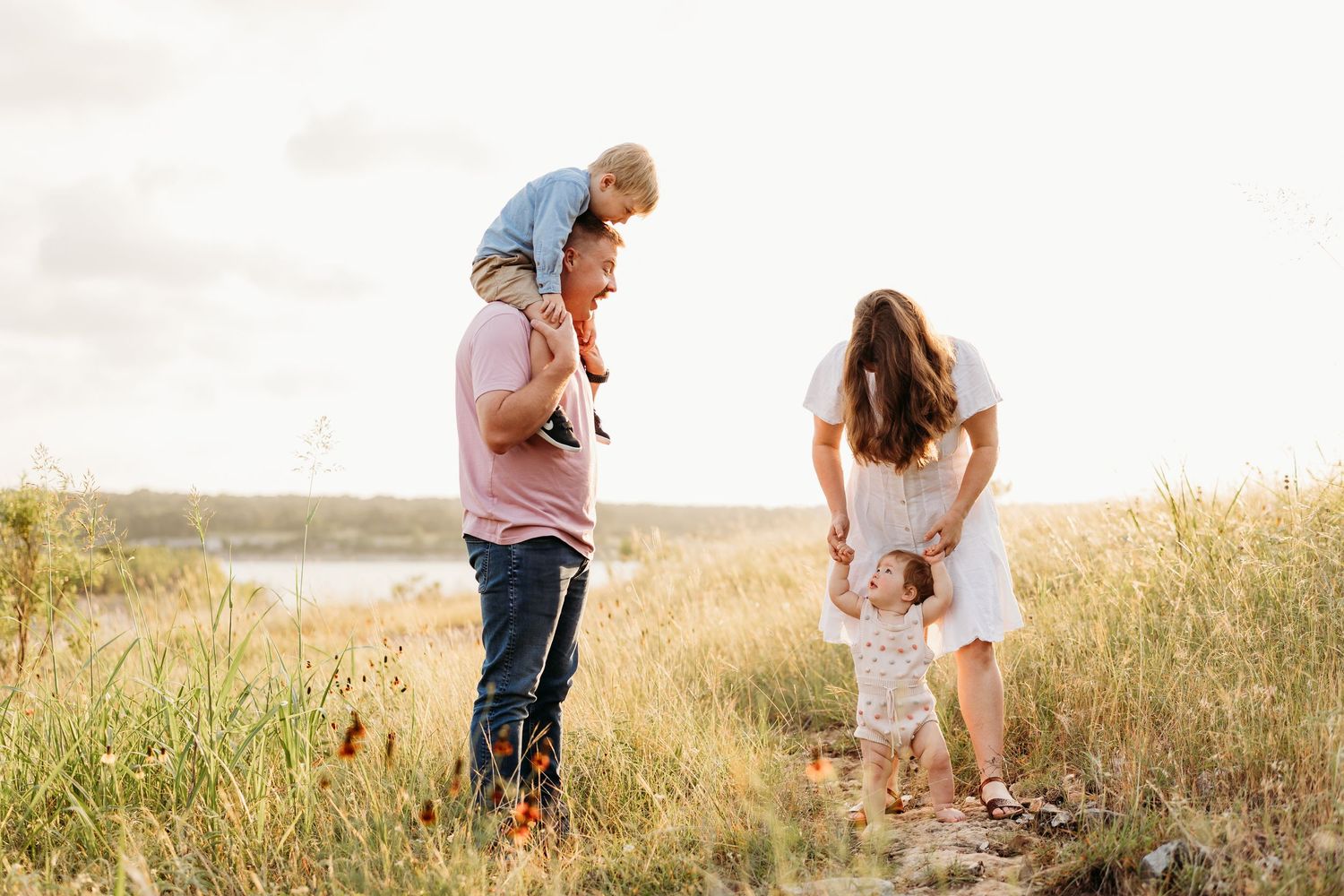 Family of four playing in golden field near water, parents with toddler on shoulders and baby girl.