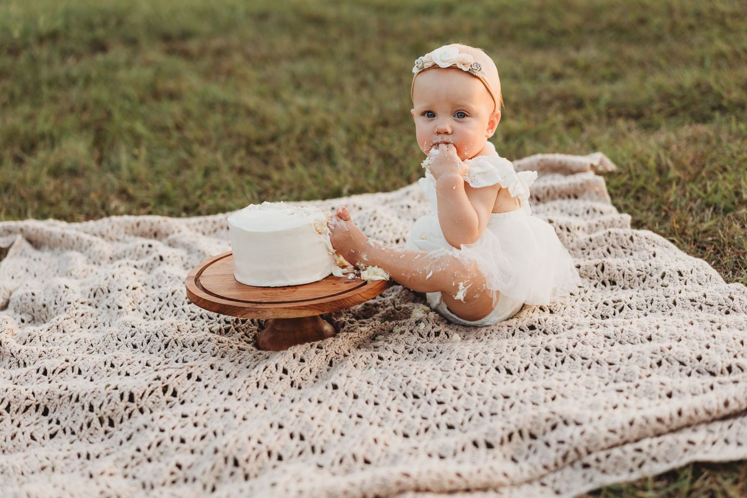 Baby girl in white tutu dress with headband sitting beside birthday cake on blanket.