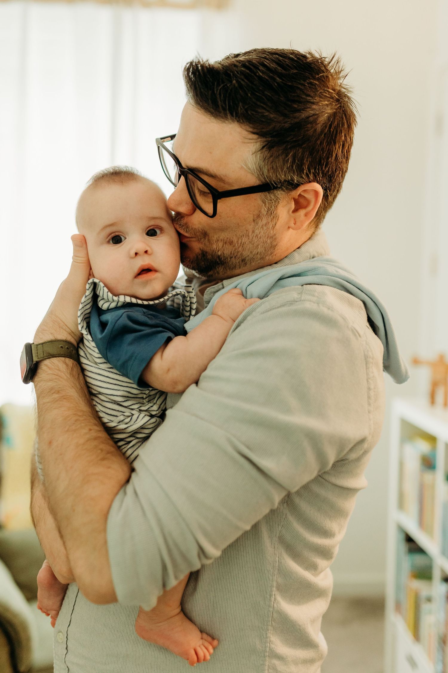 Father with glasses kissing newborn baby tenderly in bright indoor setting.
