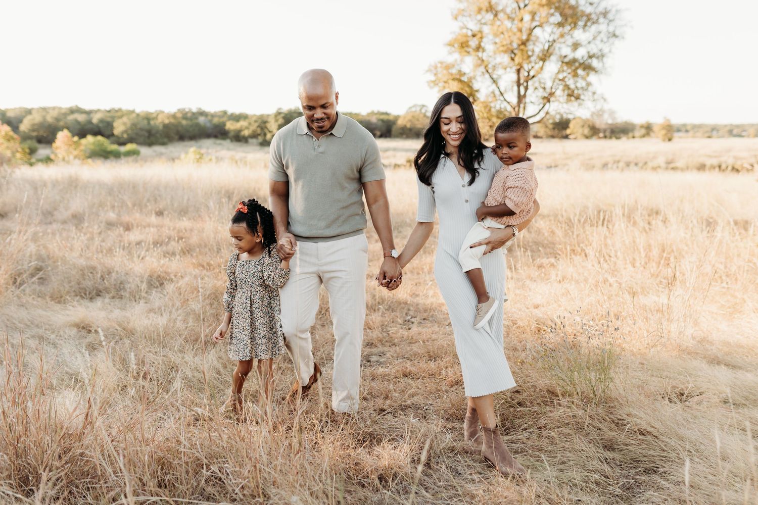 A stylish family of four walks hand in hand through a sun-drenched golden field during a portrait session.