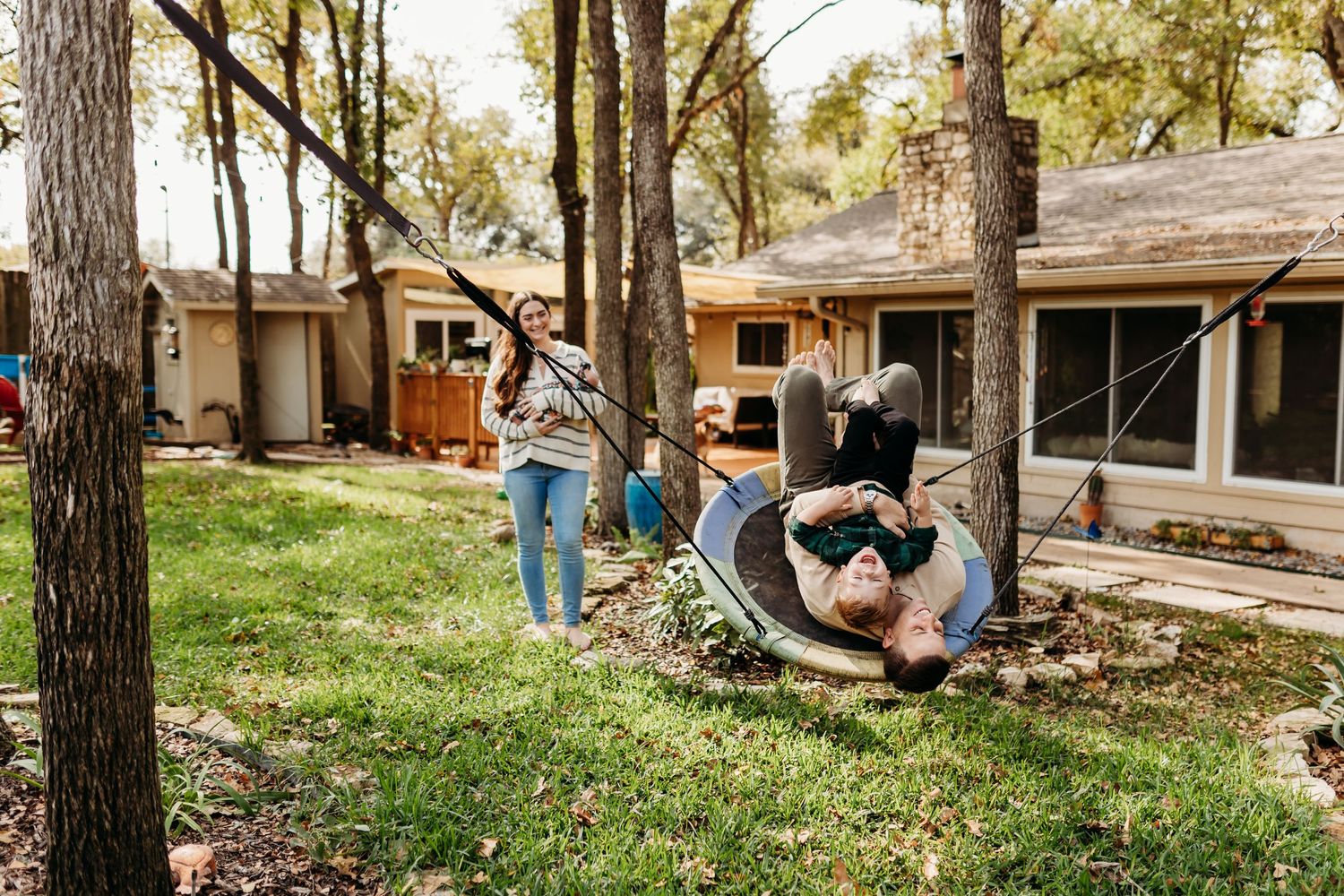Woman watching child play upside down in a hammock chair between trees in a backyard.