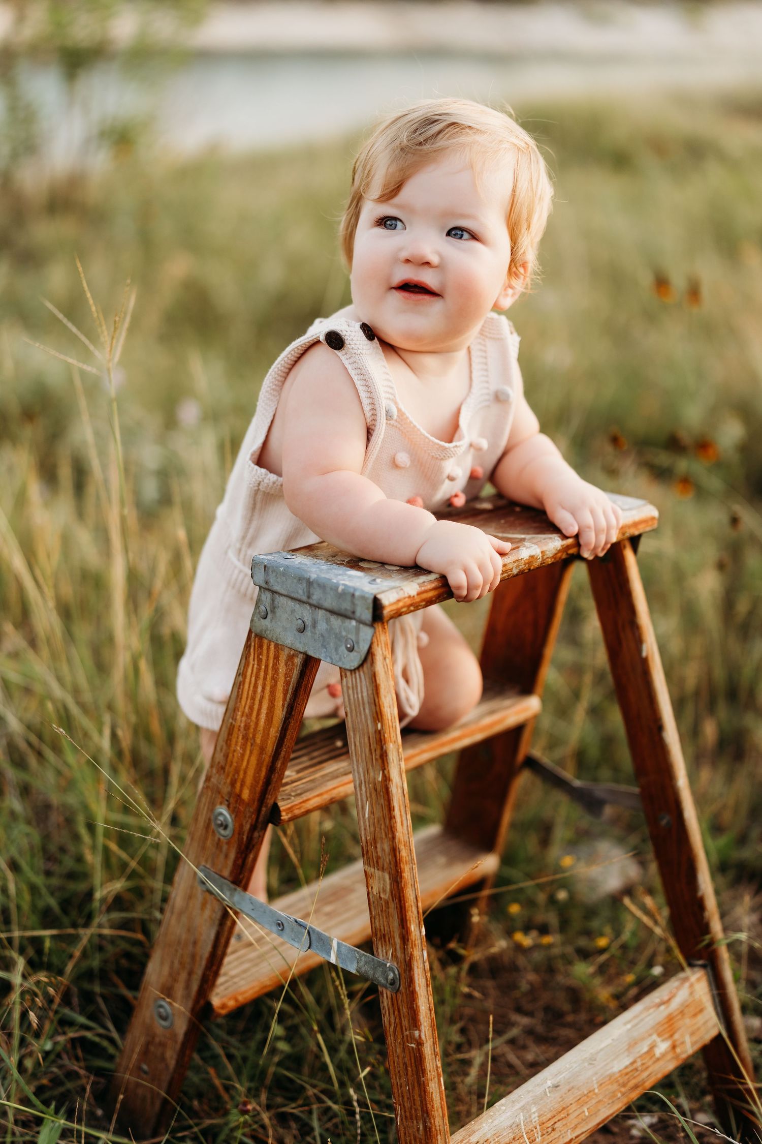 Happy baby in beige romper standing on wooden step ladder in sunny field.